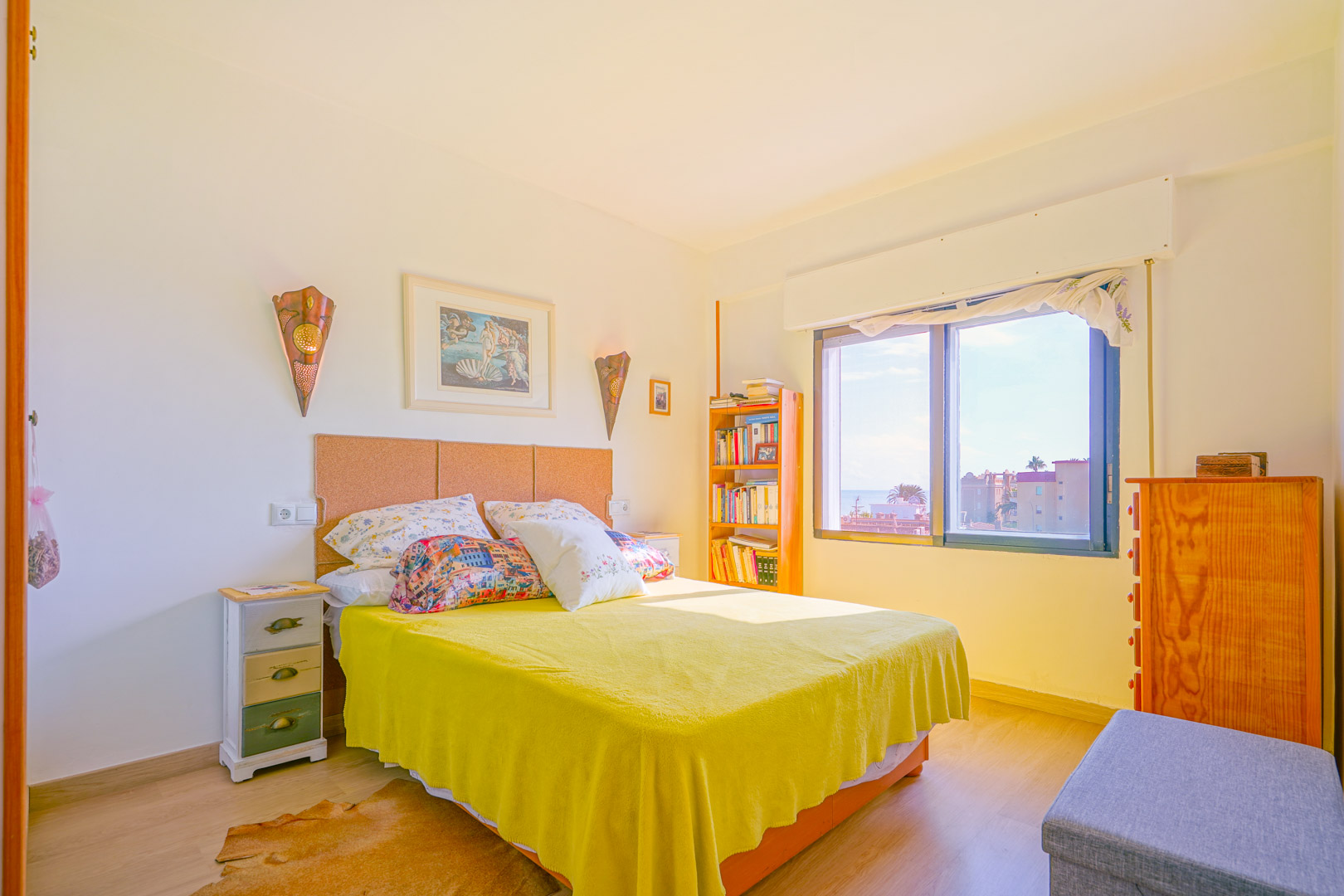 Helles Schlafzimmer mit Kork-Kopfteil, gelber Tagesdecke und Holzmöbeln. Ein großes Fenster lässt viel Tageslicht herein und bietet Ausblick auf die Stadt.