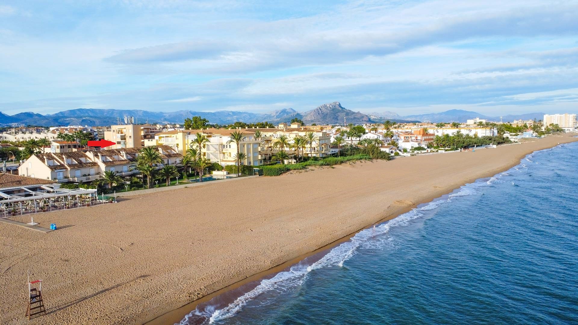 Luftaufnahme des breiten Sandstrandes und der Mittelmeerküste, die die Lage der Wohnung in Strandnähe vor der Bergkulisse zeigt.