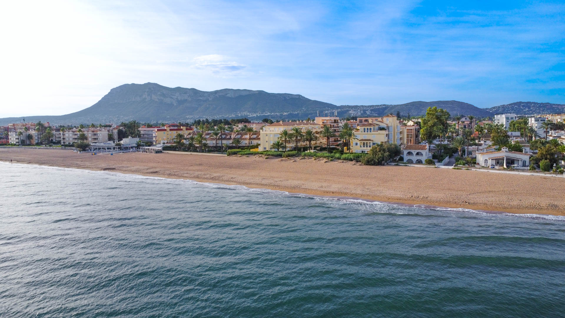 Luftaufnahme eines Sandstrandes mit Wohnanlagen und Palmen vor der Kulisse einer markanten Bergkette unter klarem blauem Himmel.