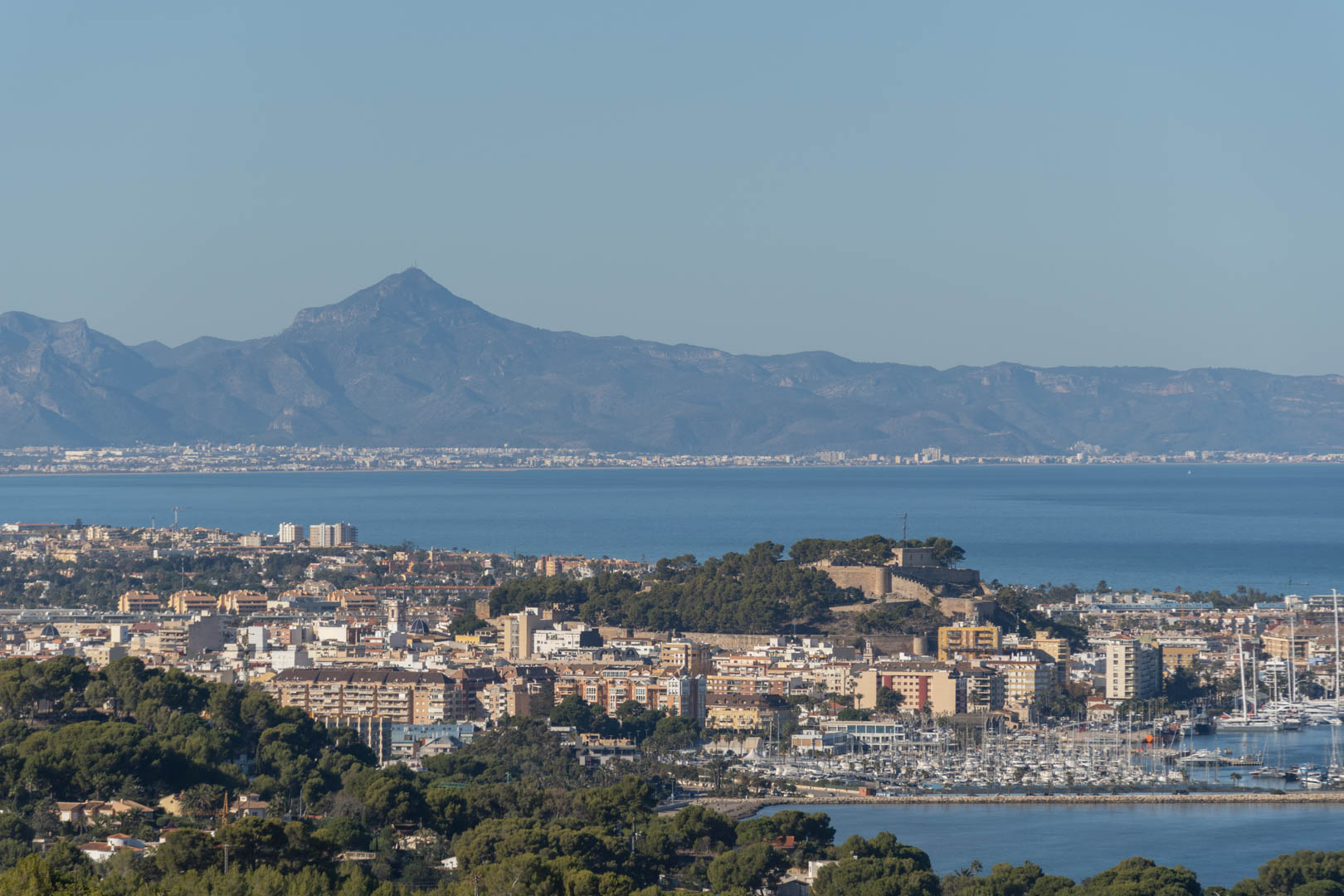 Panoramablick auf die Küstenstadt mit der historischen Burg, dem Yachthafen und einer markanten Bergkette im Hintergrund über der Mittelmeerbucht.