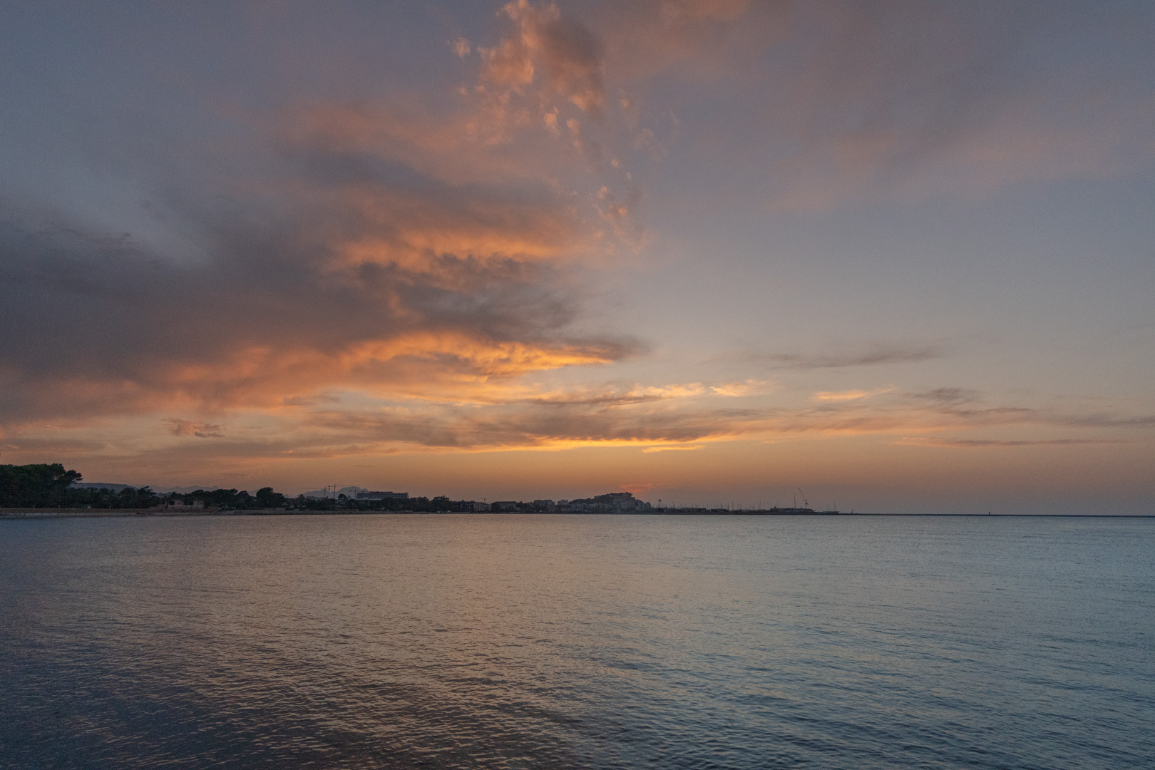 Panoramablick auf die Mittelmeerküste bei Sonnenuntergang mit orangefarbenen Wolken und ruhigem Wasser, das den Abendhimmel über der fernen Stadt widerspiegelt.