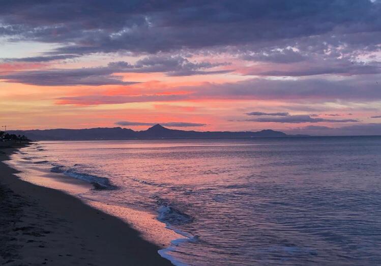 Panoramablick auf die Sandküste in der Abenddämmerung mit Bergsilhouetten unter einem violett-orangen Himmel.