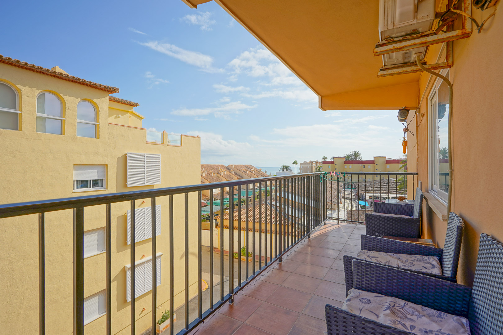 Privater Balkon mit Terrakotta-Fliesen, schwarzem Metallgeländer und Korbsesseln. Er bietet Ausblicke auf die umliegende Architektur und den fernen Meeresspiegel.