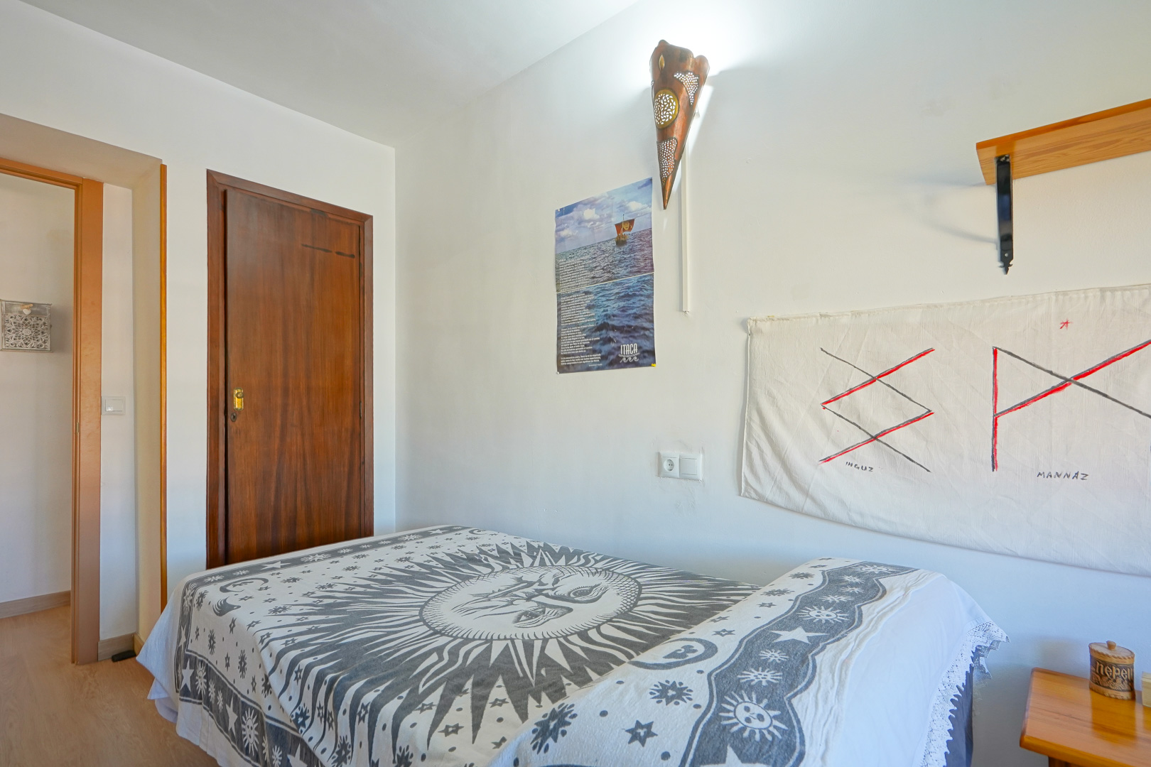 Schlafzimmer mit Einzelbett und Tagesdecke mit Sonnenmotiv, weißen Wänden mit verzierter Kupferwandleuchte und einer dunklen Massivholztür.