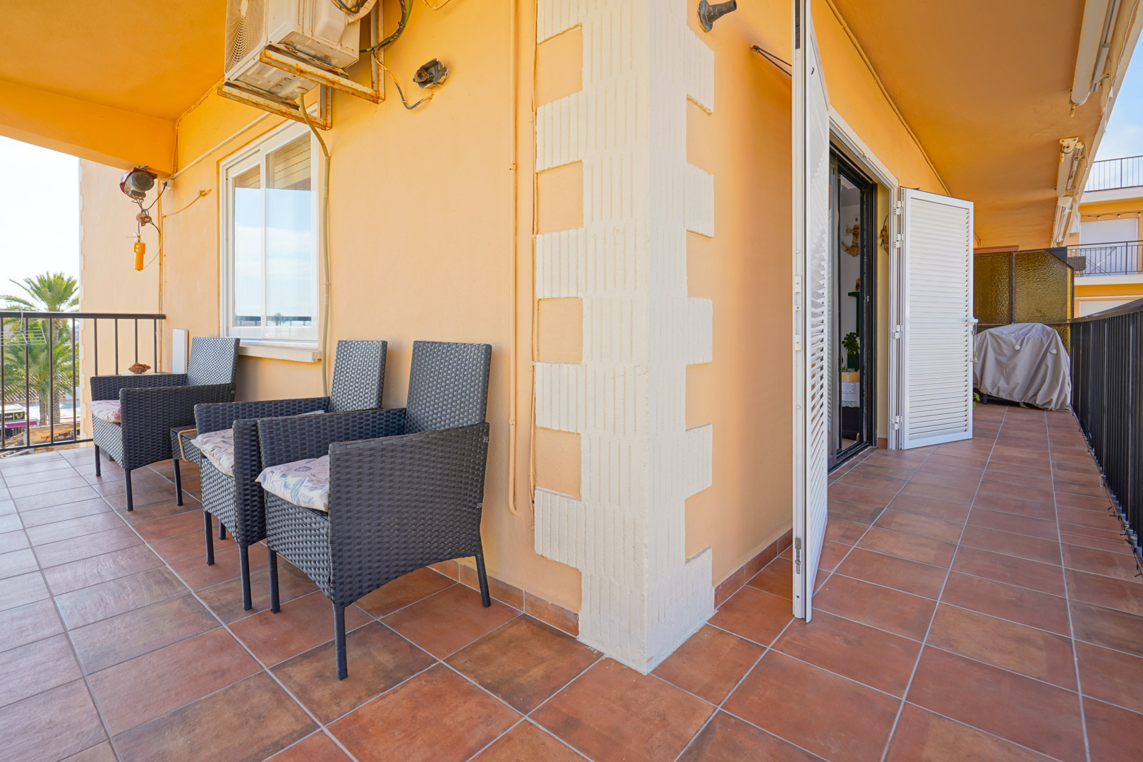 Überdachte Terrasse mit Terrakottaboden, dunklen Rattansesseln und weißen Fensterläden. Die Wand ist mit einer Klimaanlage und dekorativen Säulenelementen versehen.