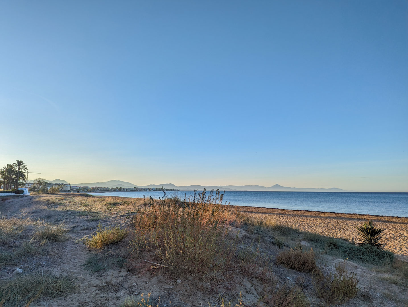 Weite Aussicht auf einen naturbelassenen Strand mit Dünen und Wildwuchs, mit Blick auf das ruhige Meer und eine Bergkette am Horizont.