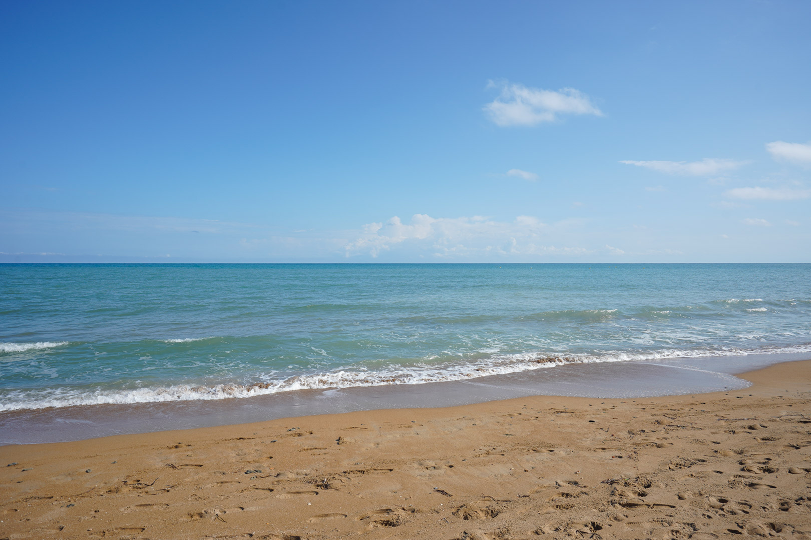 Weiter Sandstrand mit sanften türkisfarbenen Wellen unter einem klaren blauen Himmel. Das Ufer besticht durch goldenen Sand und ruhiges Meereswasser.