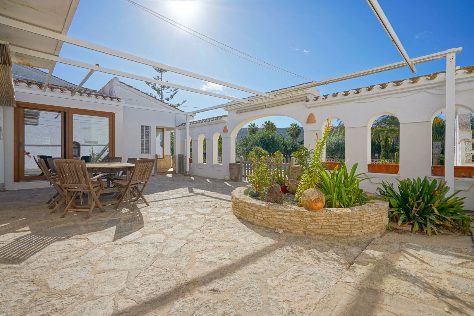 Außenanlage mit Natursteinpflaster, Essbereich aus Holz und rundem Steinbeet unter einer Metall-Pergola-Konstruktion.