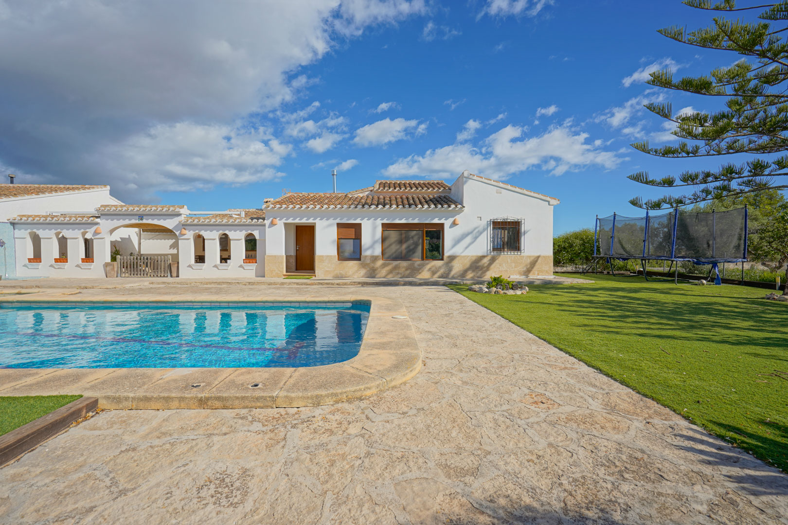 Außenansicht der Villa mit großem Pool, Natursteinterrasse und gepflegtem Rasen, die den mediterranen Baustil mit weißen Wänden und Ziegeldach zeigt.