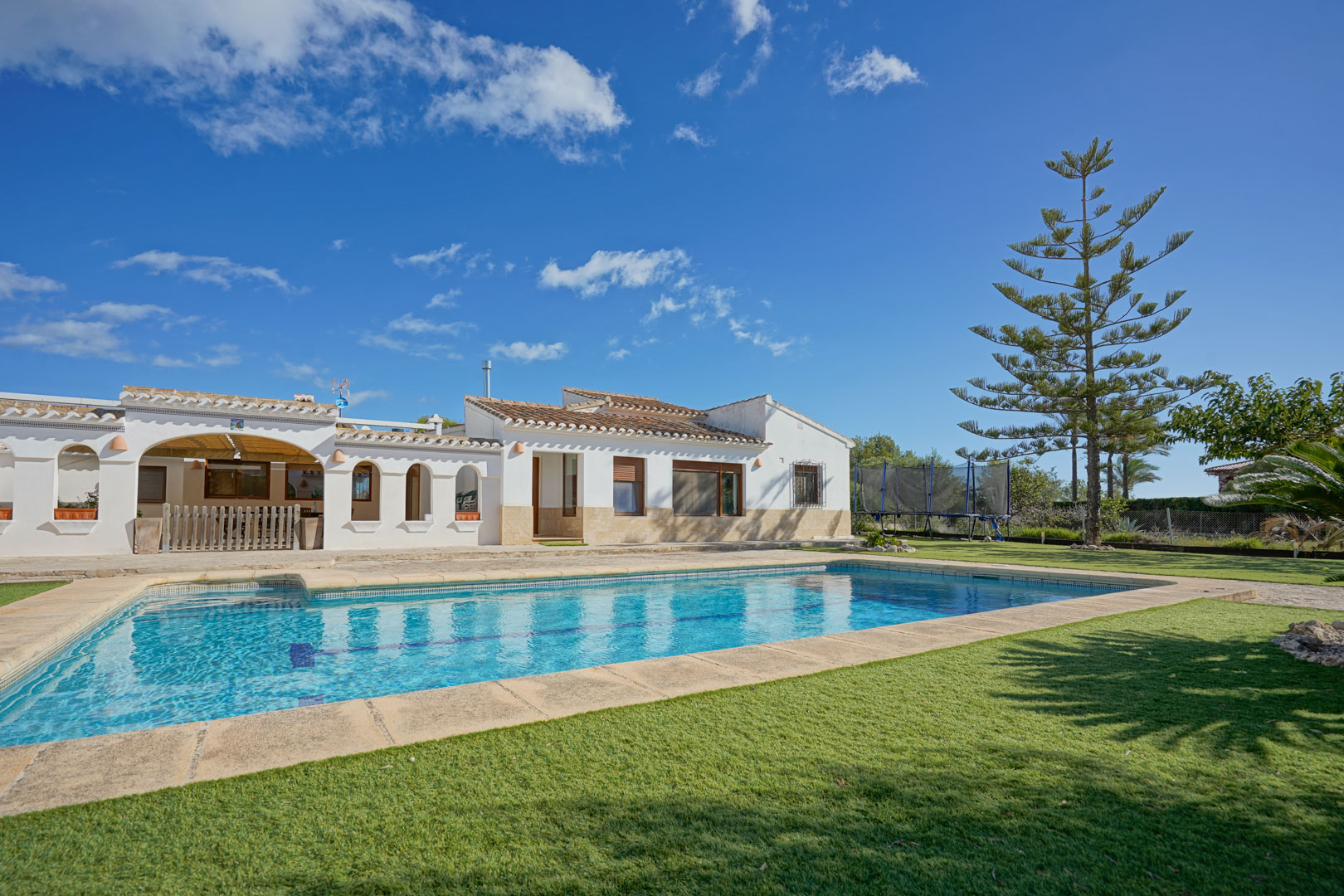 Außenansicht der Villa mit rechteckigem Pool, gepflegtem Rasen und Steinterrasse. Die weiße Fassade besticht durch traditionelle Rundbögen und ein klassisches Ziegeldach.