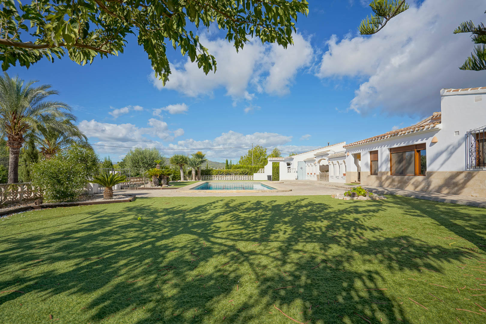 Außenansicht der weißen Villa mit Steinsockel, rechteckigem Pool, gepflasterter Sonnenterrasse und gepflegtem Garten mit Rasenfläche und Palmen.