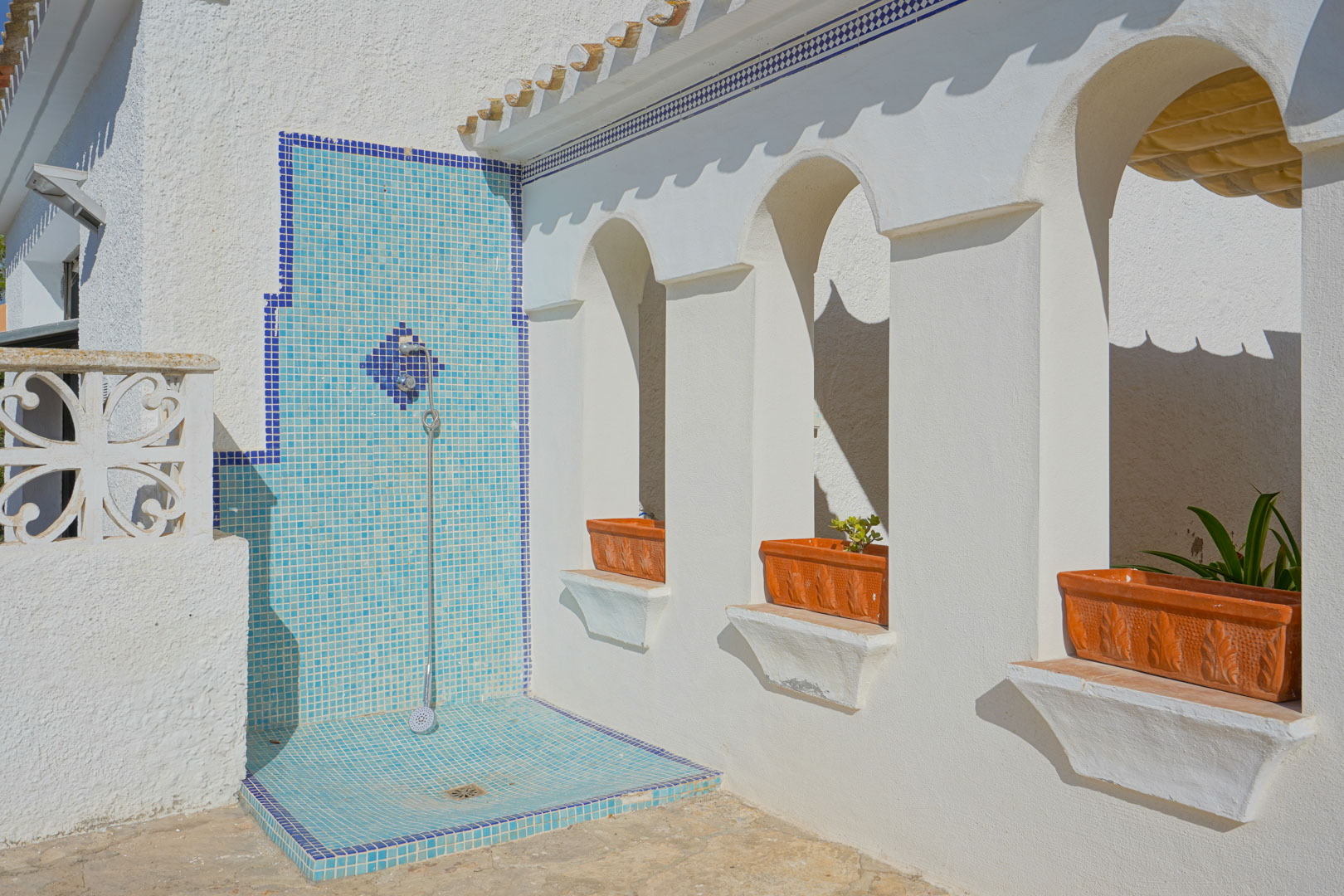 Außendusche mit blauem Mosaikfliesen-Dekor neben einer weißen Fassade mit Rundbögen und integrierten Terrakotta-Pflanzgefäßen.