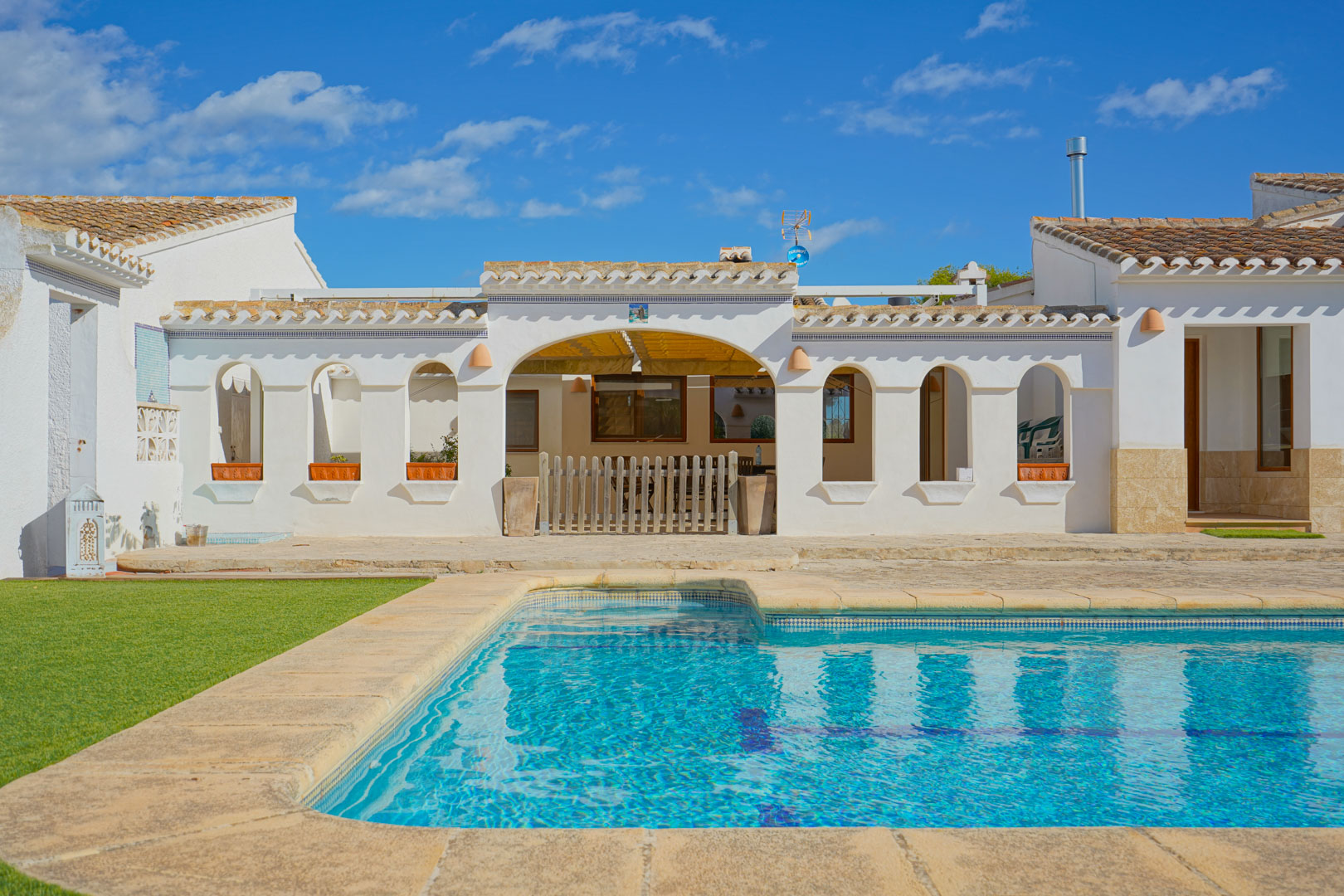 Außenpool mit Natursteineinfassung und Sonnenterrasse vor einer weißen mediterranen Fassade mit traditionellen Rundbögen und Terrakotta-Ziegeldach.