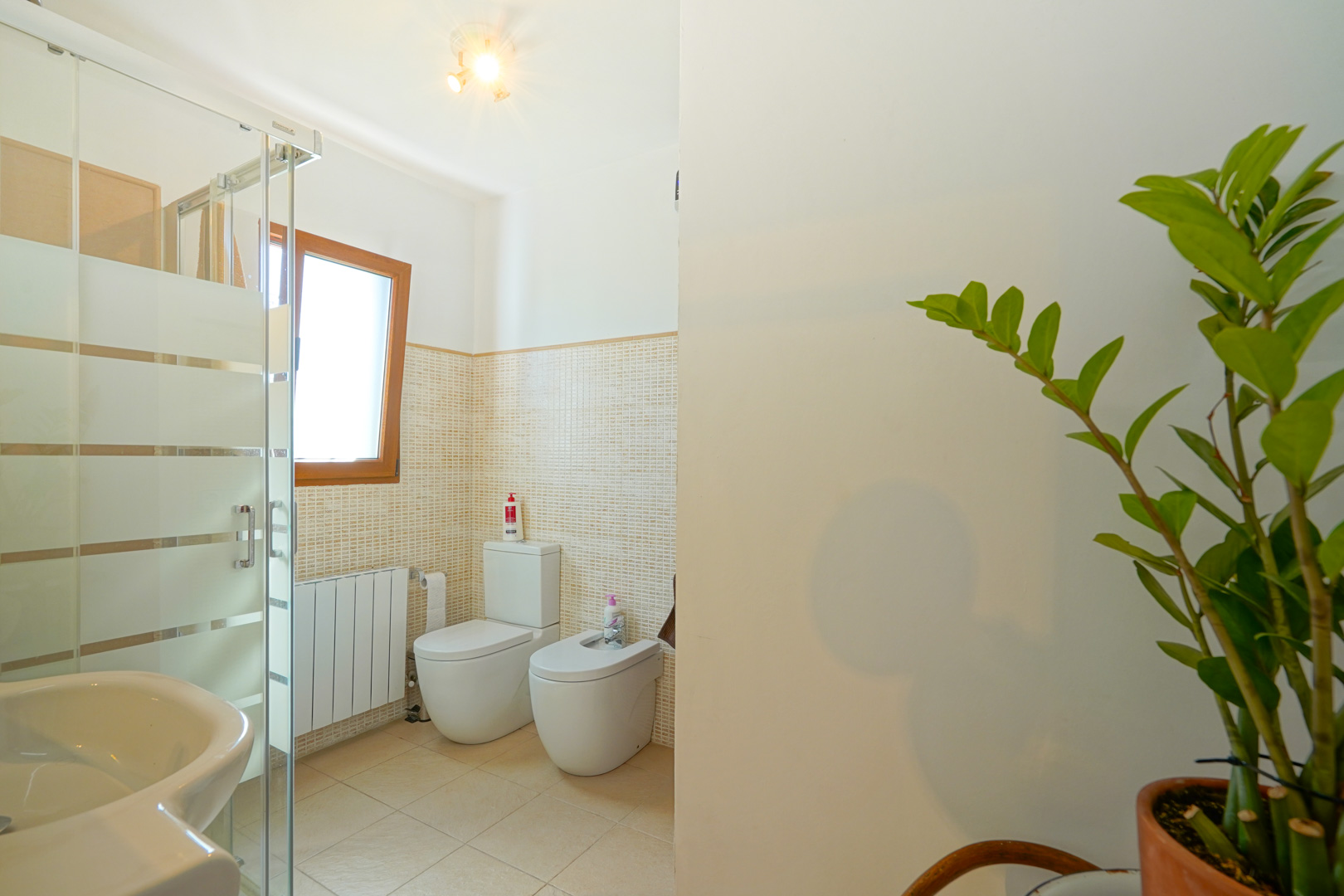 Badezimmer mit Glasduschkabine, WC, Bidet und Heizkörper, ausgestattet mit beigefarbenen Mosaikfliesen und einem Fenster mit Holzrahmen.