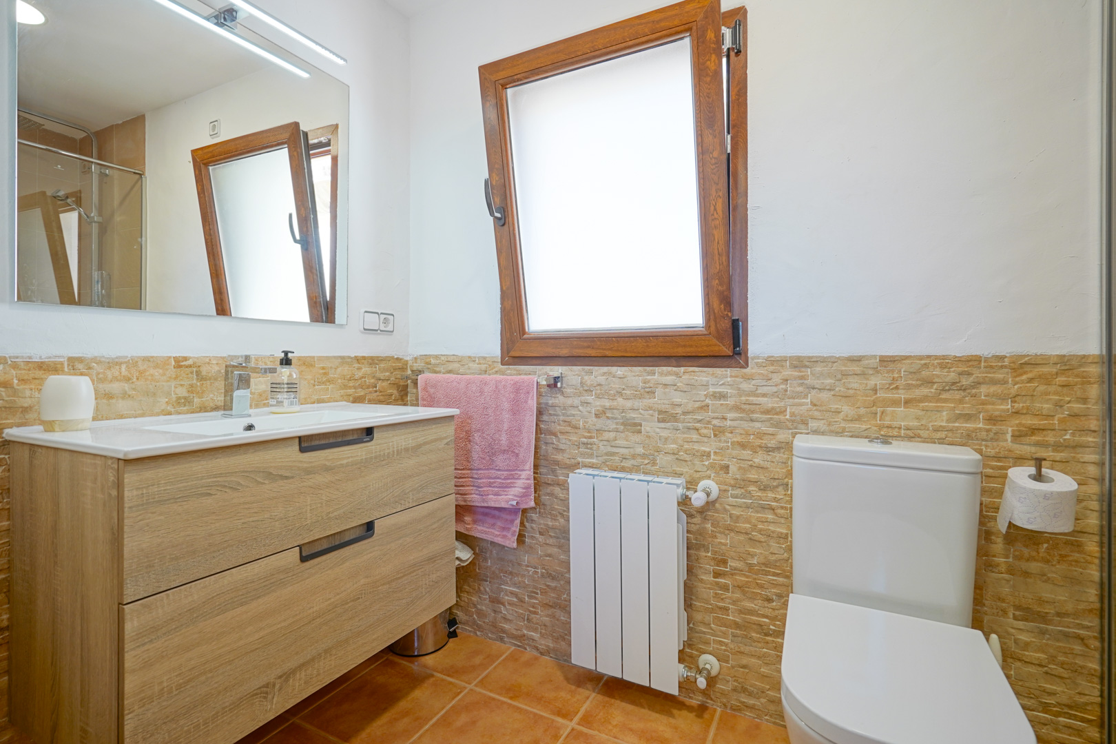 Badezimmer mit Holzwaschtisch, LED-beleuchtetem Spiegel und Wandfliesen in Steinoptik. Ausgestattet mit einem Heizkörper und einem Dreh-Kipp-Fenster aus Holz.