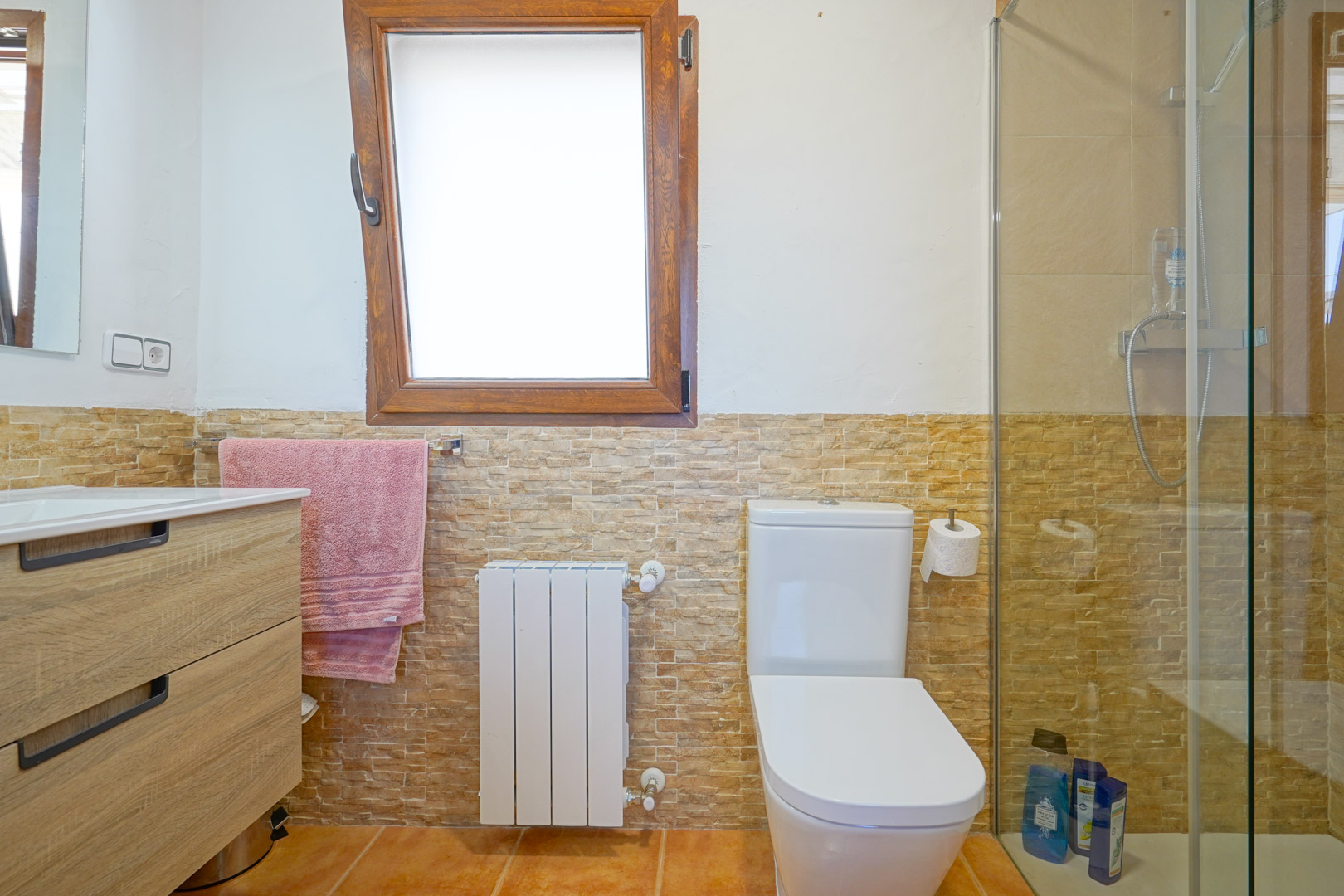 Badezimmer mit Waschtisch in Holzoptik, Steinmosaik-Wandfliesen, weißem Heizkörper und einer begehbaren Dusche mit Glastrennwand neben einem Holzfenster.