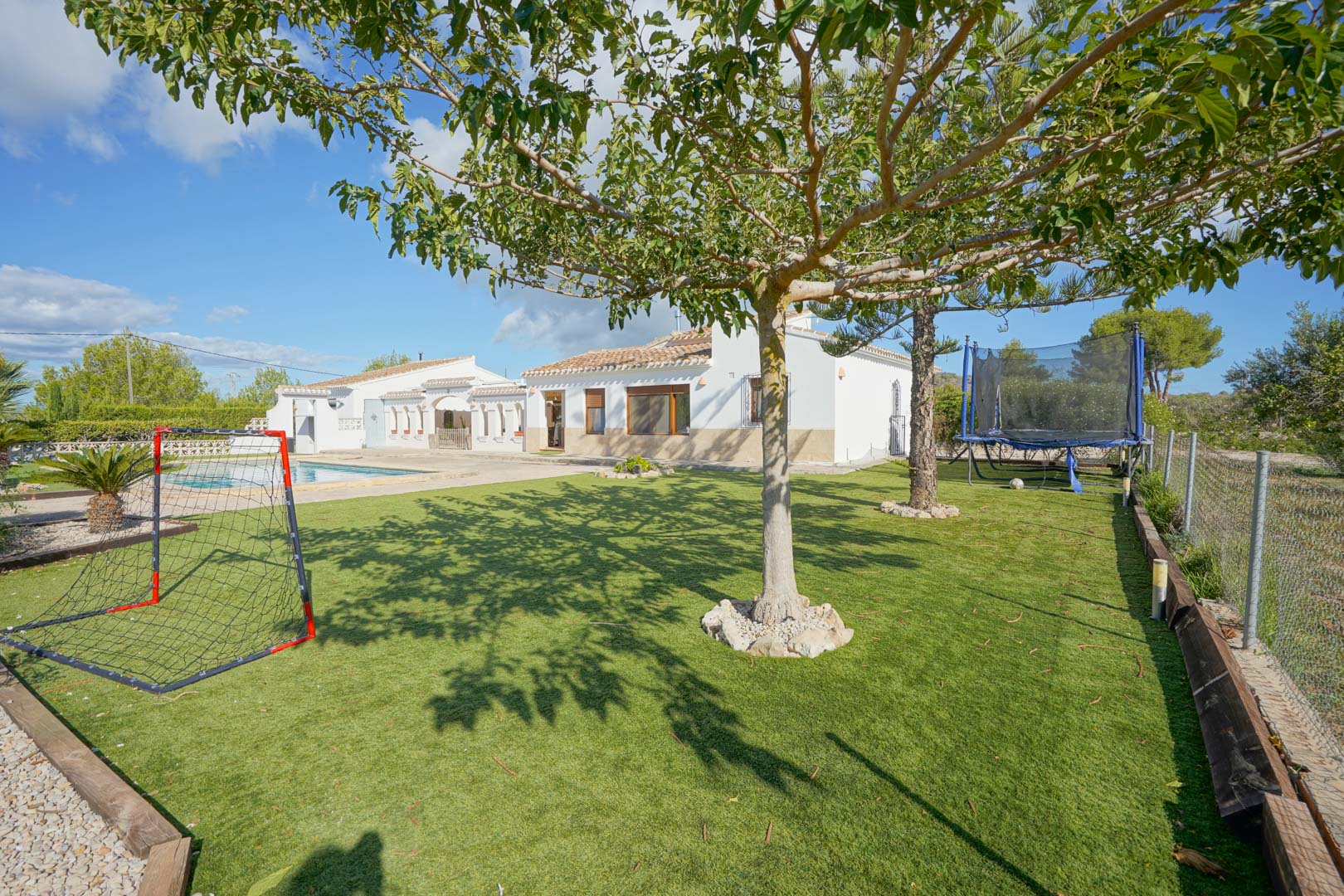 Großzügiger Garten mit Kunstrasen und Freizeitbereich inklusive Fußballtor und Trampolin, gelegen vor der weißen Fassade der Villa und dem Pool.