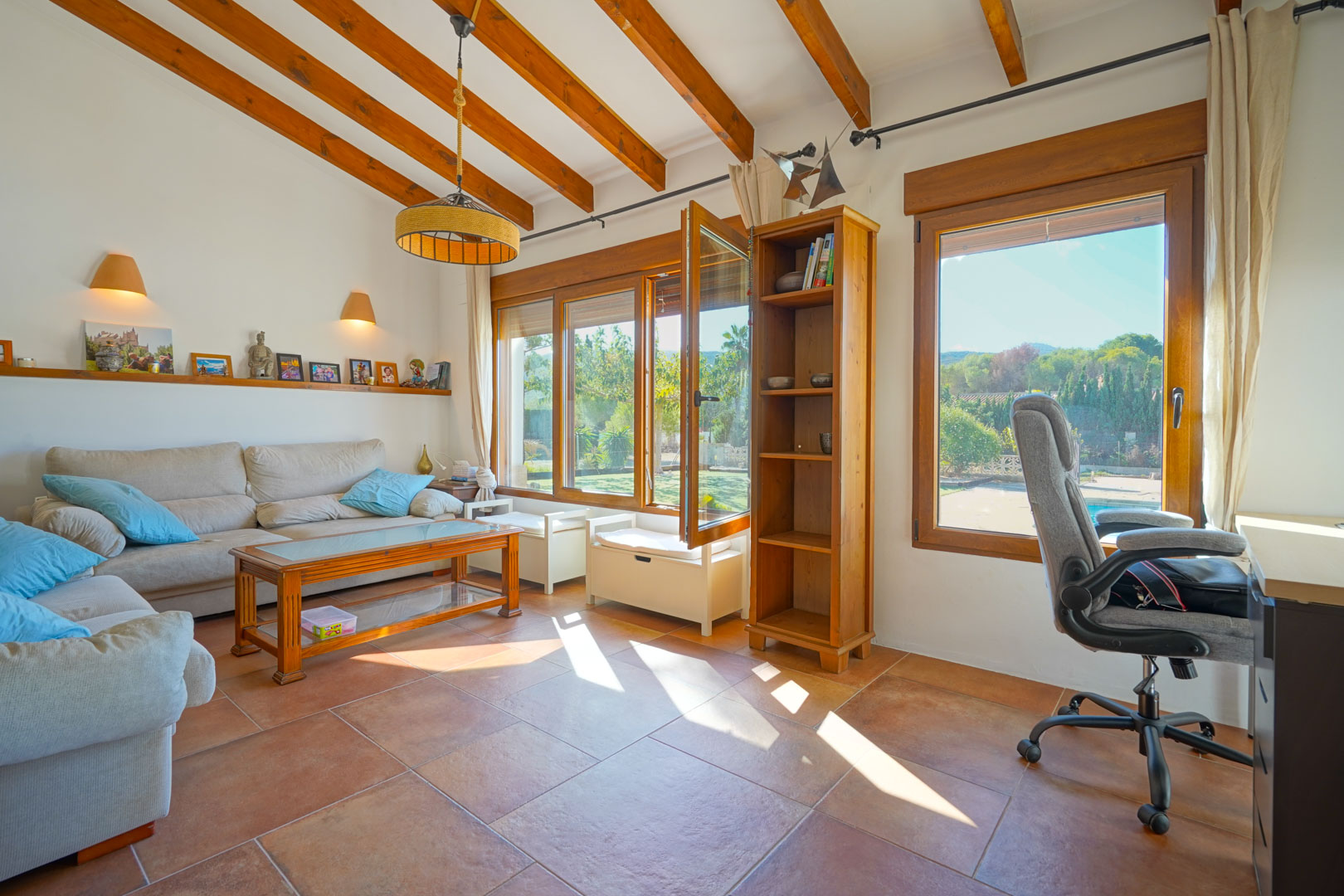 Heller Wohnraum mit Holzbalkendecke, Terrakottafliesen und integriertem Büro. Große Holzfenster bieten einen direkten Ausblick auf den Garten und den Pool.