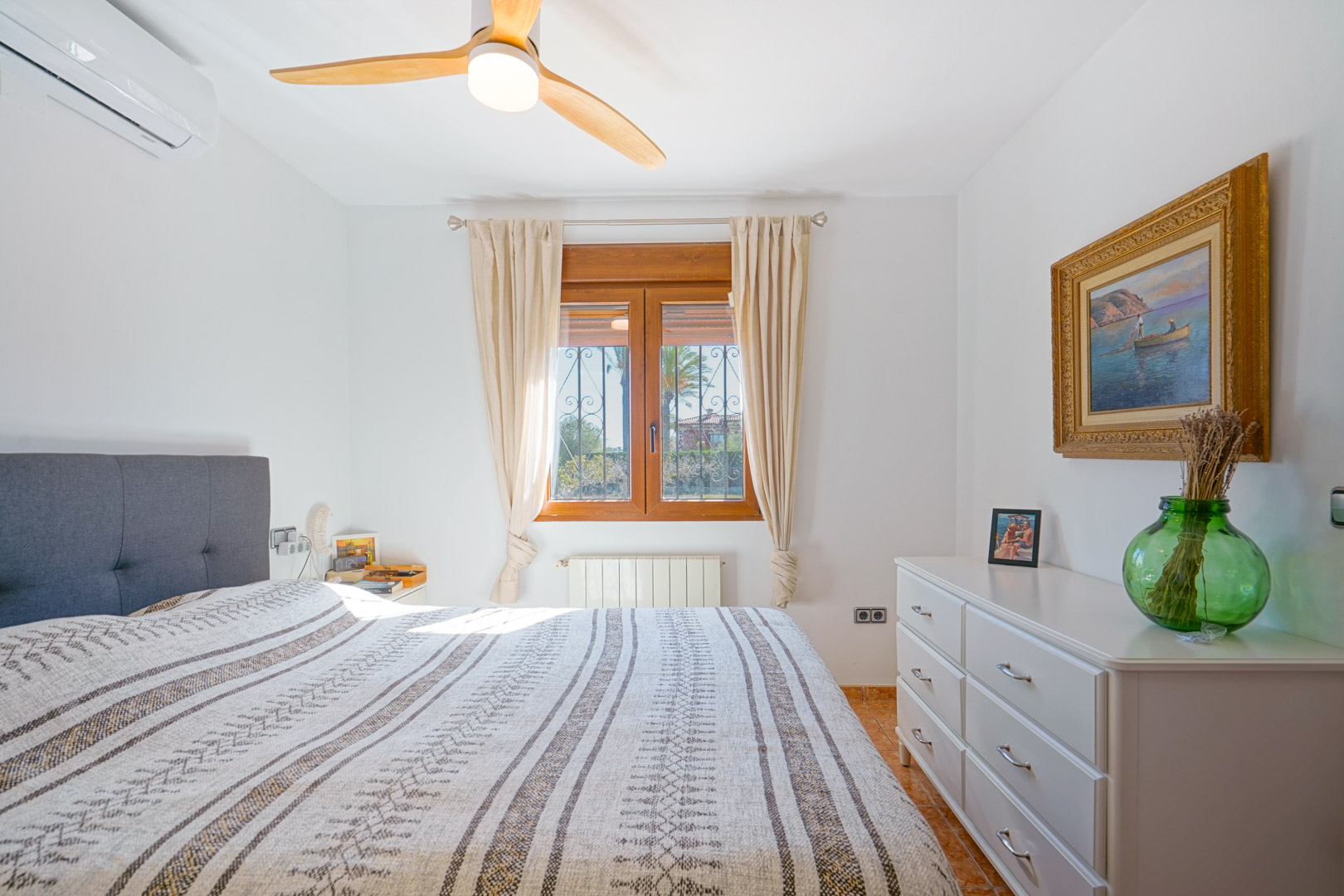 Helles Schlafzimmer mit Holzfensterrahmen, Deckenventilator mit Holzflügeln, Klimaanlage und weißer Kommode. Ausgestattet mit Heizkörper und Fliesenboden.