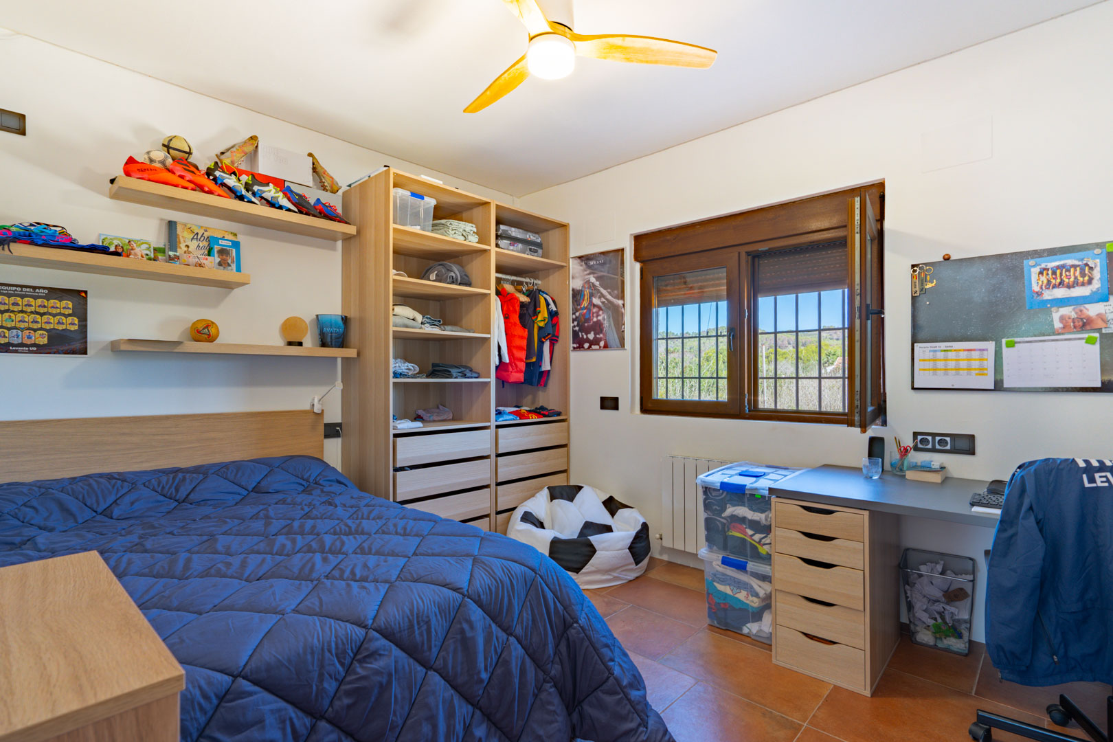 Helles Schlafzimmer mit Terrakottaboden, integriertem Schreibtisch und offenem Holzregalsystem. Ein Holzfenster bietet Ausblick auf die umliegende, grüne Landschaft.
