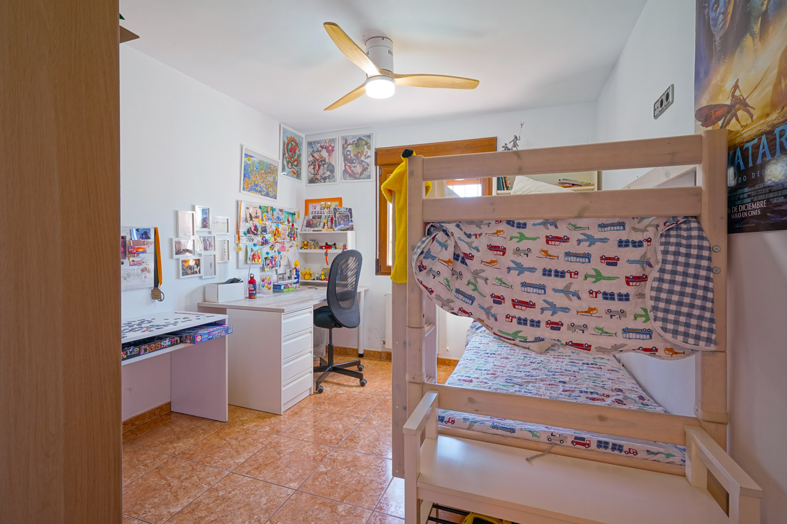 Kinderzimmer mit Etagenbett aus Holz, Schreibtisch und Deckenventilator. Der Raum ist mit Fliesenboden ausgestattet und bietet Tageslicht durch ein Fenster mit Holzrahmen.