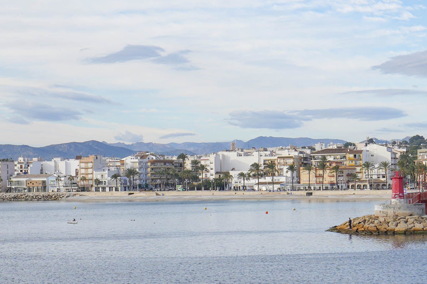 Panoramablick auf die Küste von Jávea mit dem markanten roten Leuchtturm, Gebäuden am Wasser und Bergen im Hintergrund.