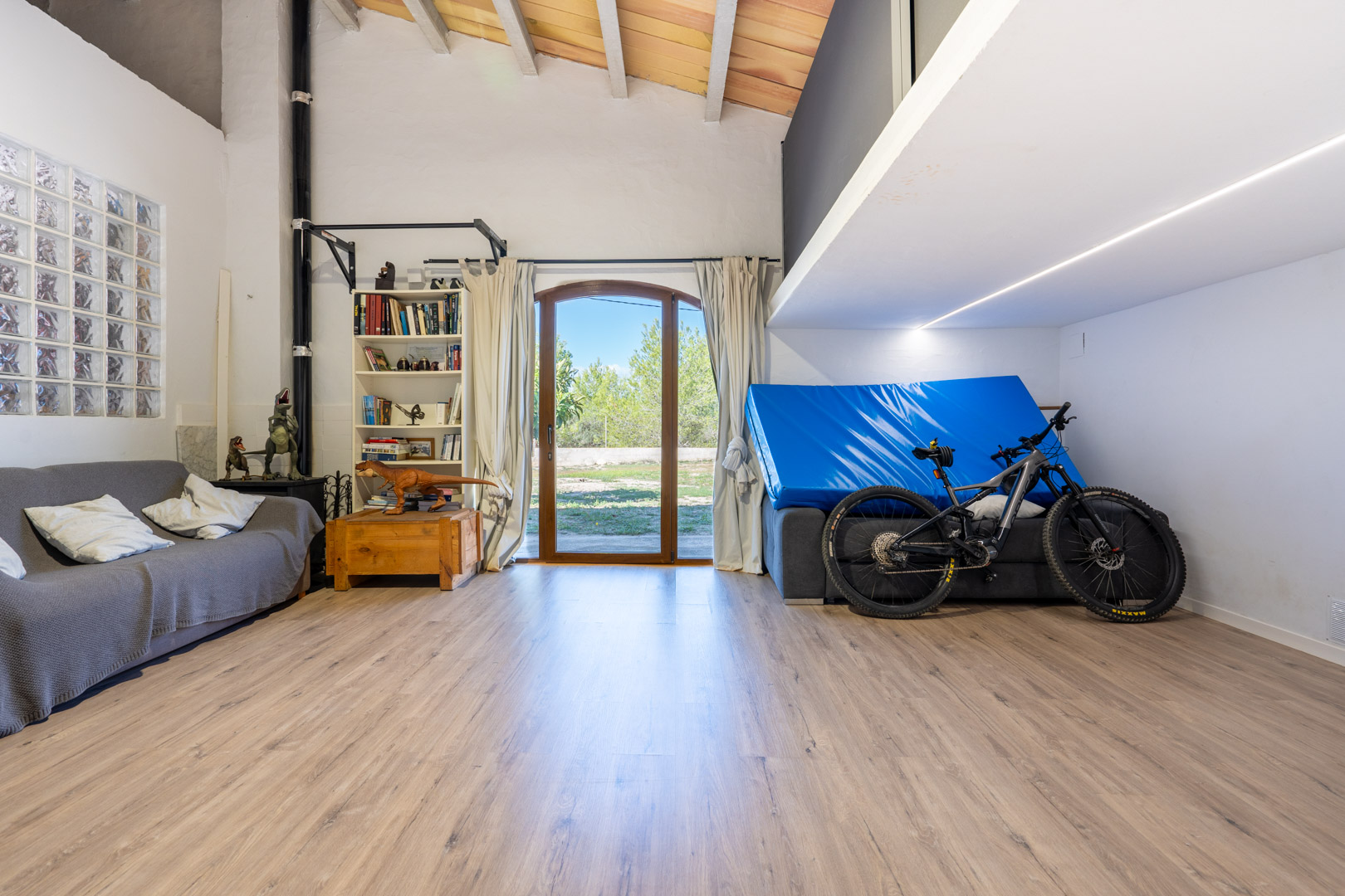 Vielseitig nutzbarer Raum mit hohen Decken, sichtbaren Holzbalken, einer Galerieebene, Laminatboden und direktem Zugang zum Garten durch eine Rundbogentür.