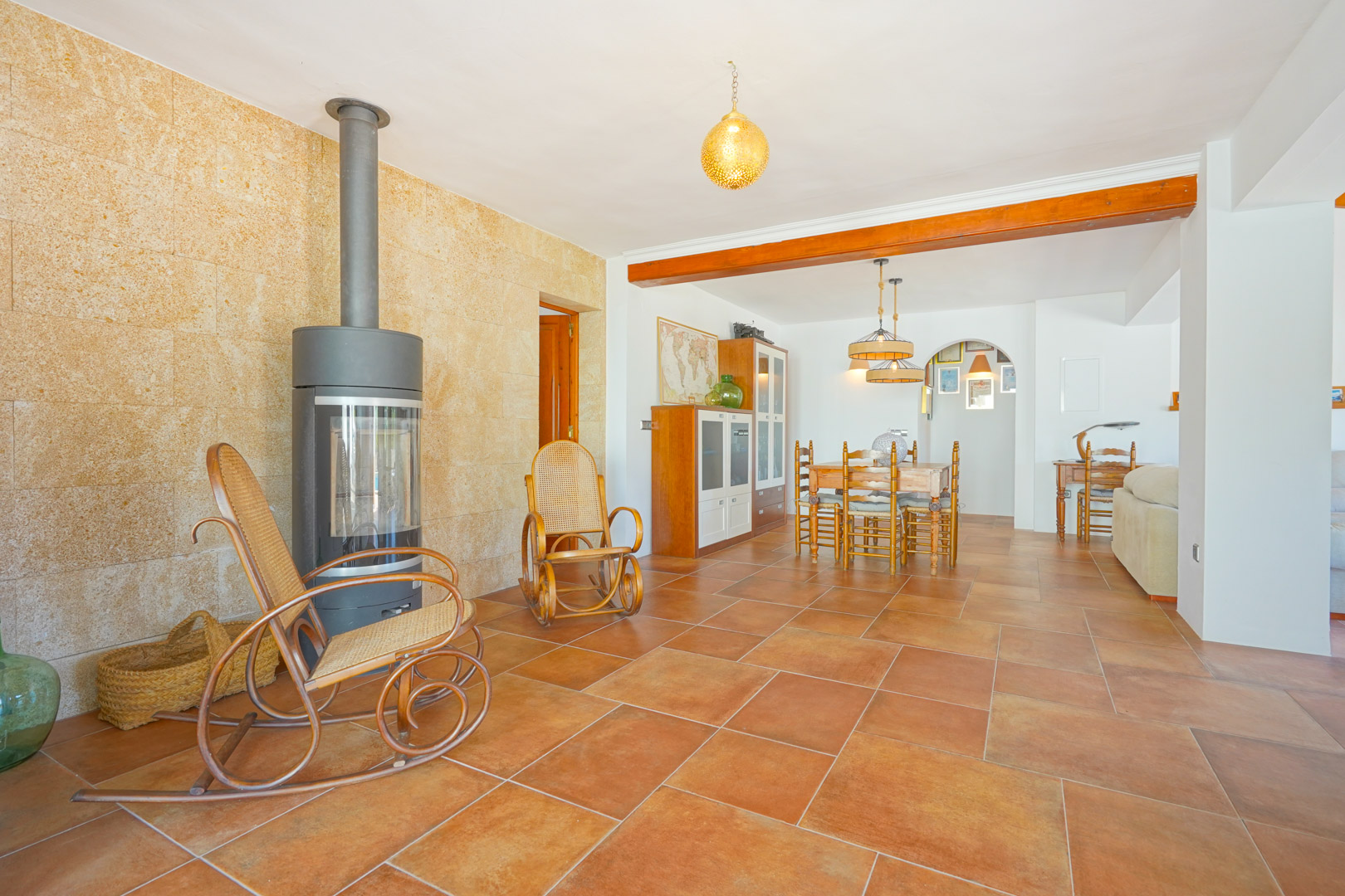 Wohnbereich mit modernem Kaminofen vor einer Natursteinwand, Terrakottaböden und integriertem Essplatz unter einem Sichtbalken aus Holz.