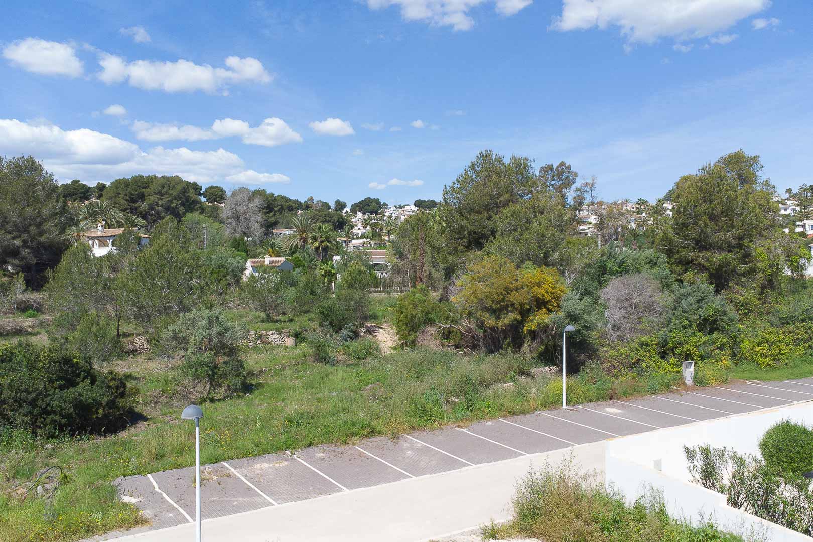 Panoramablick vom Anwesen auf die mediterrane Landschaft mit üppiger Vegetation, umliegenden Häusern und einem Außenparkplatz unter blauem Himmel.