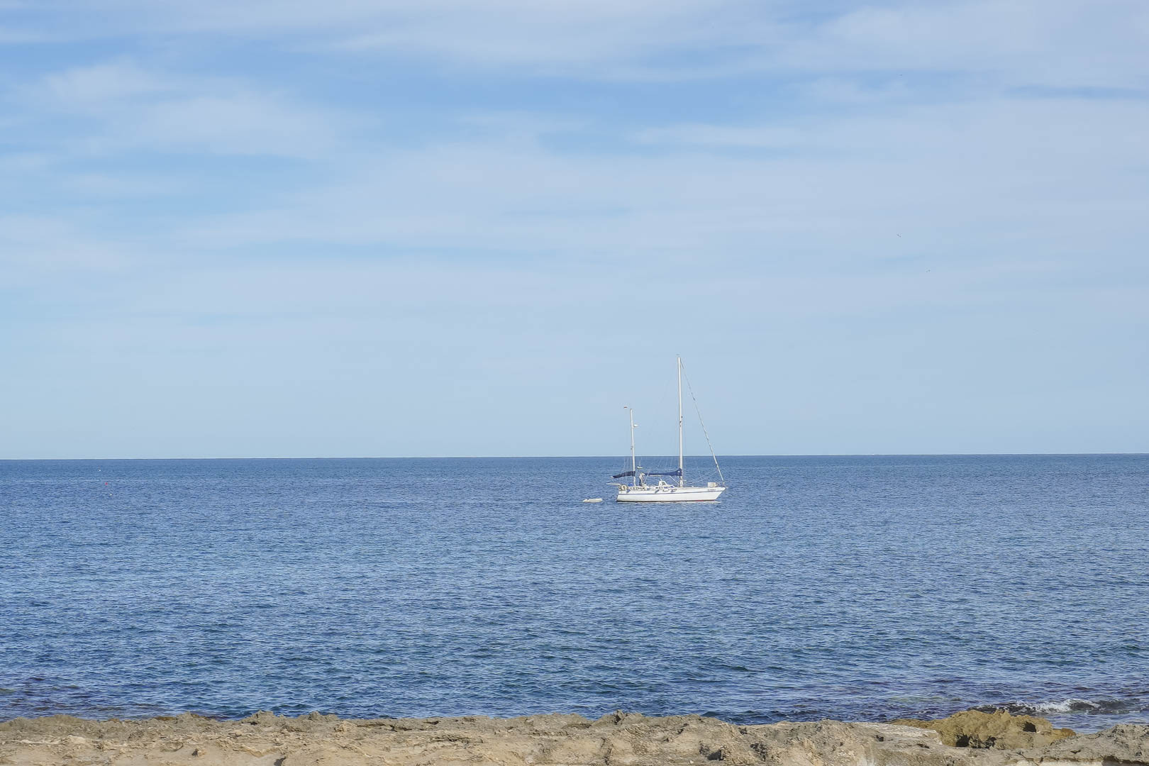 Panoramablick auf das Mittelmeer von der Küste von Jávea aus, mit einem Segelboot vor der felsigen Küste unter klarem Himmel.