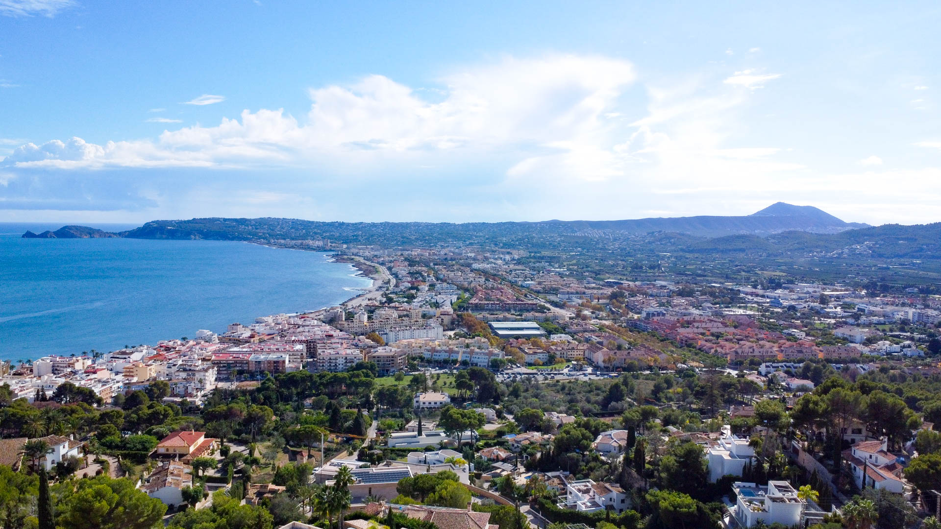 Panoramablick aus der Vogelperspektive auf die Bucht von Jávea und den Hafen, mit der Mittelmeerküste und dem Montgó-Massiv im Hintergrund.