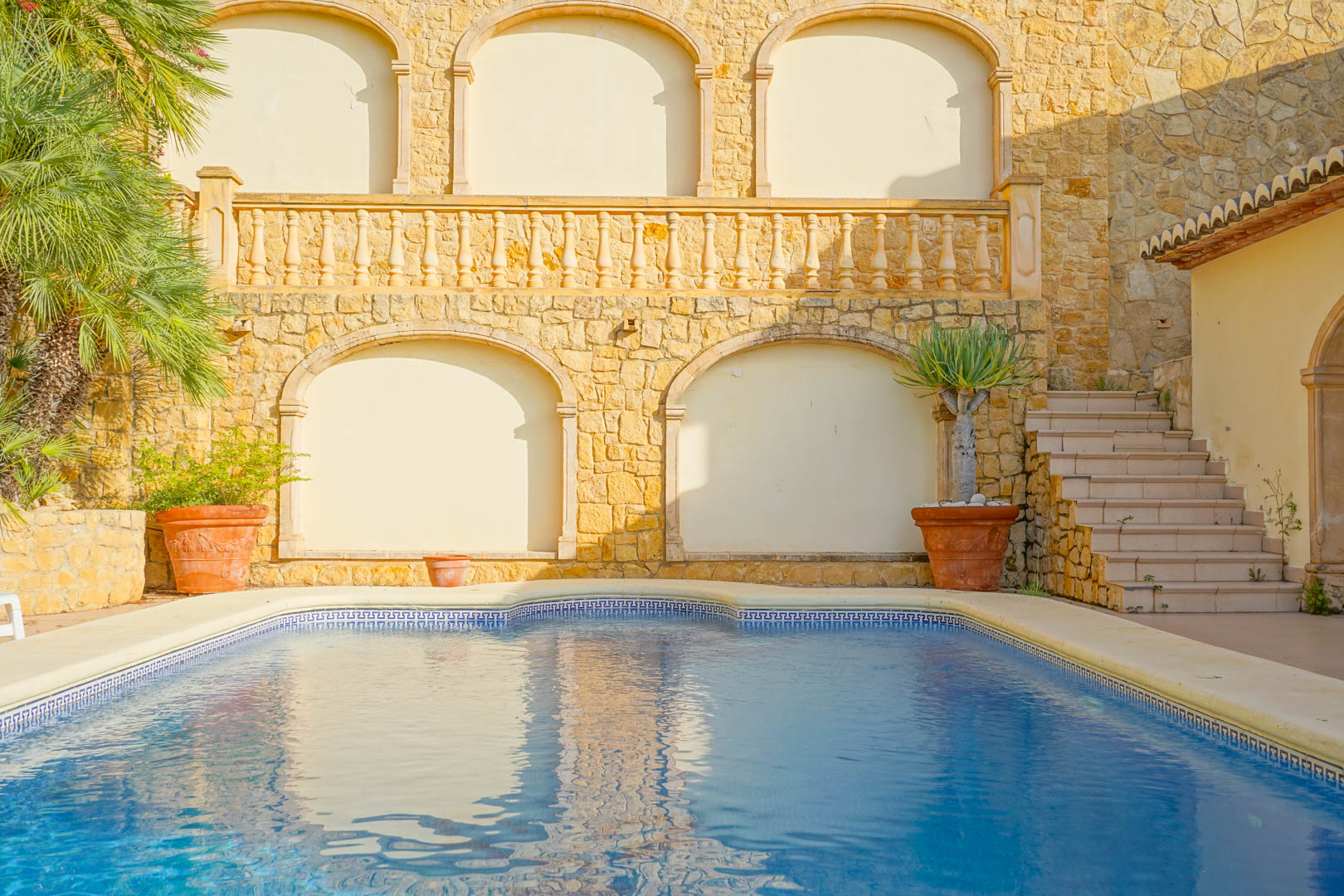 Außenpool mit blauen Mosaikfliesen und griechischer Borte, umgeben von Natursteinmauern mit Rundbogennischen, einer klassischen Balustrade und mediterranen Pflanzen.