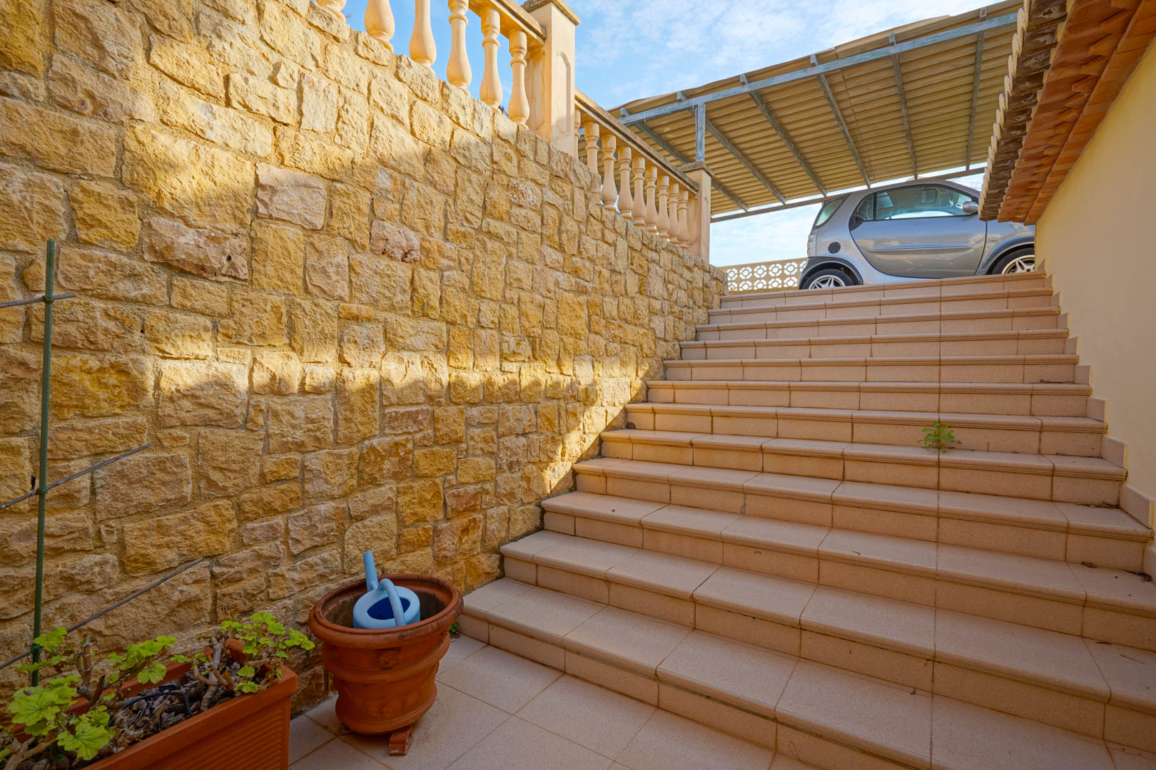 Außentreppe mit Keramikfliesen und einer Natursteinmauer, die die Wohnebenen mit dem überdachten Carport auf der oberen Ebene verbindet.