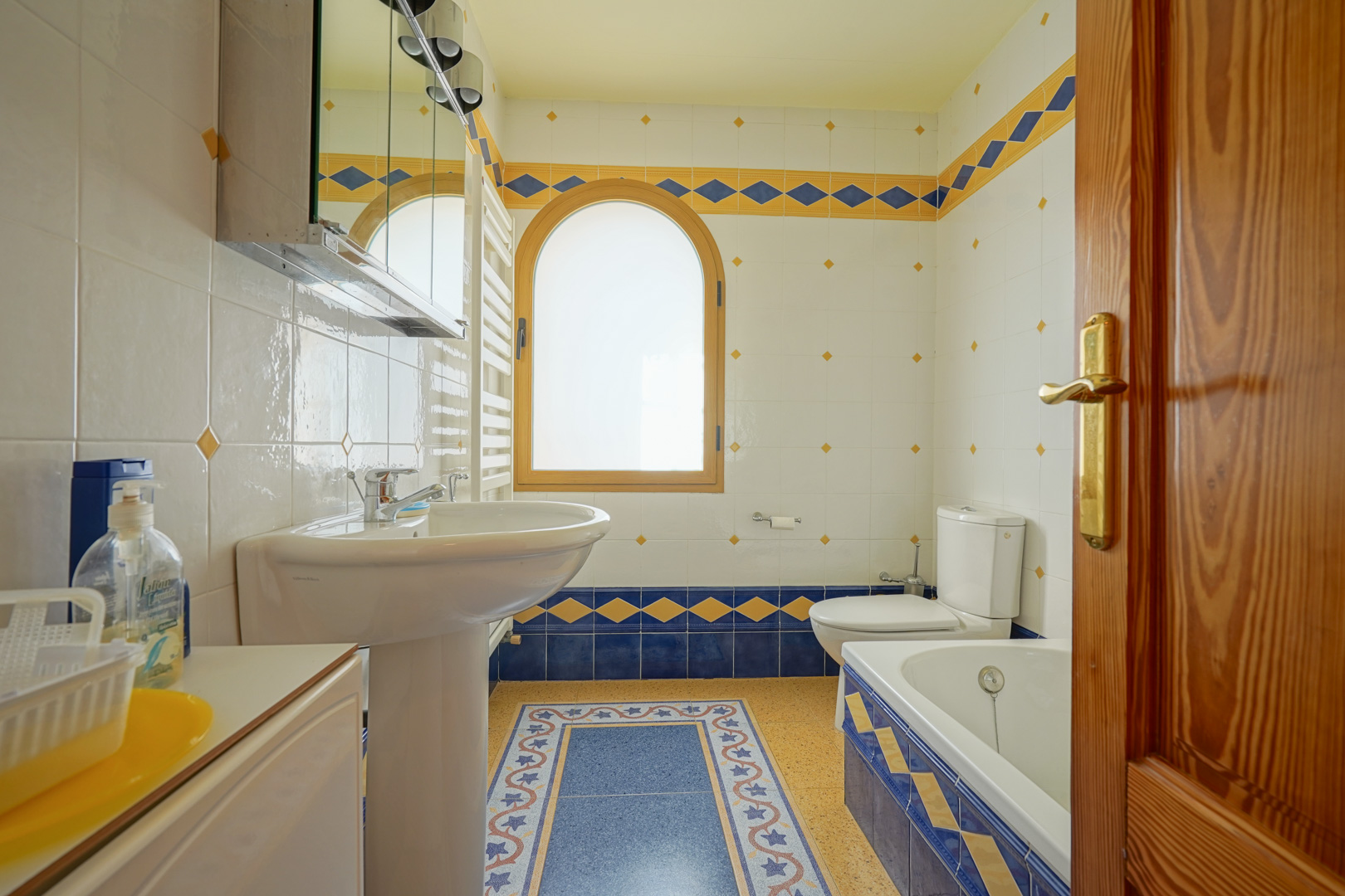 Badezimmer mit dekorativen blau-gelben Fliesen, einem Rundbogenfenster aus Holz sowie einer Ausstattung mit Badewanne, Säulenwaschbecken und Handtuchheizkörper.