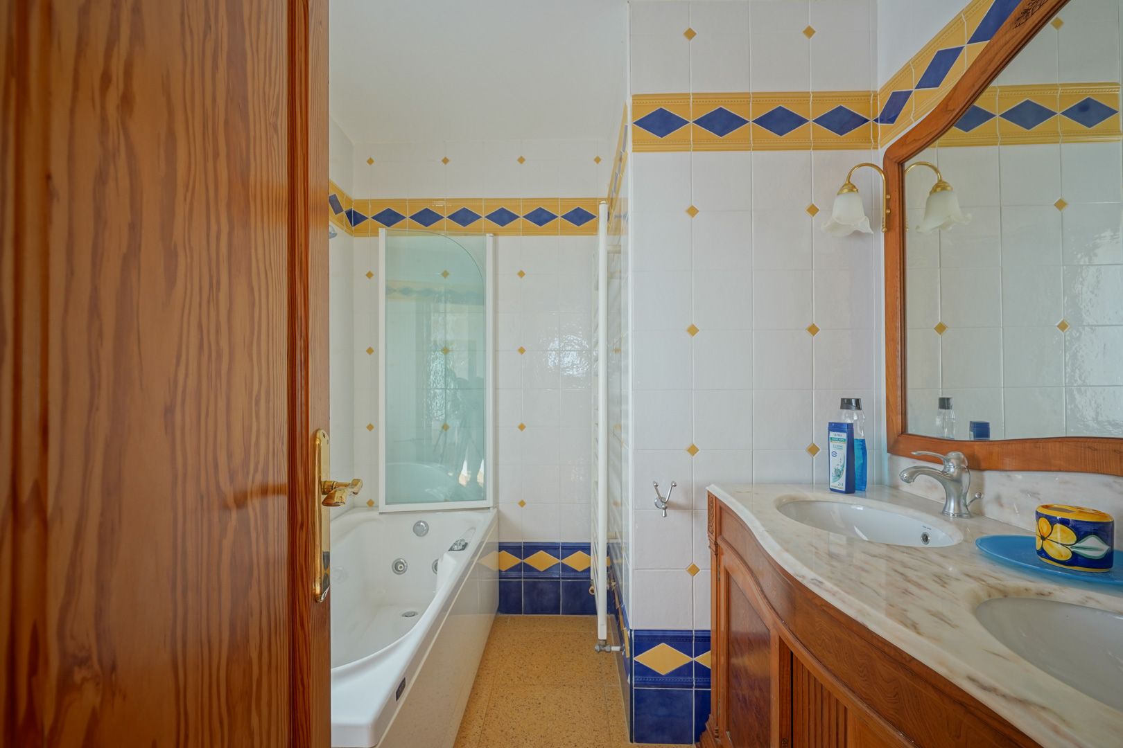 Badezimmer mit Doppelwaschtisch aus Marmor, Holzunterschrank und Hydromassage-Badewanne, akzentuiert durch blau-gelbe Fliesenbordüren im mediterranen Stil.