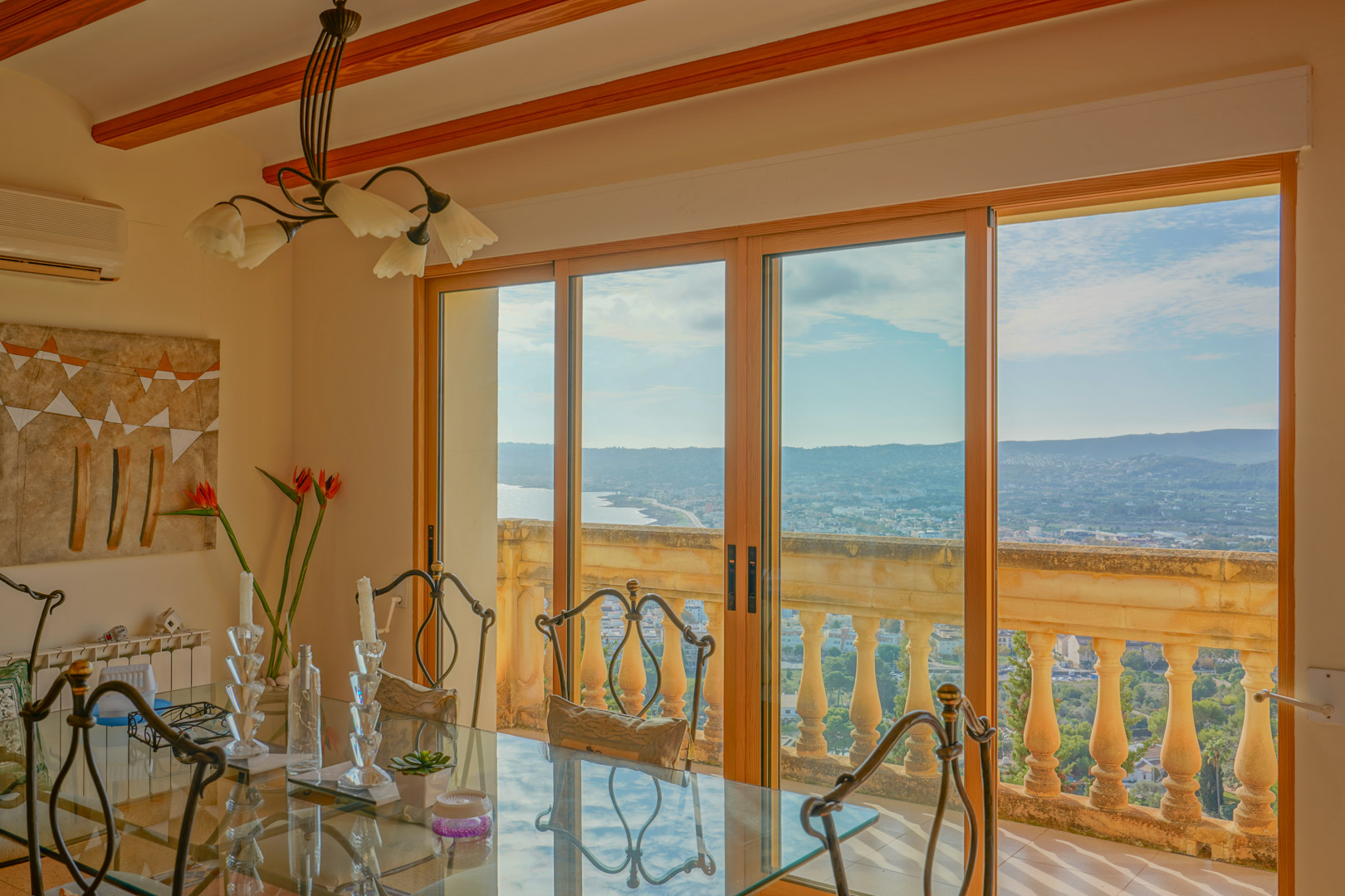Essbereich mit Glastisch und Holzbalkendecke, der durch große Schiebetüren einen weiten Blick auf die Küste von Jávea und das Mittelmeer bietet.