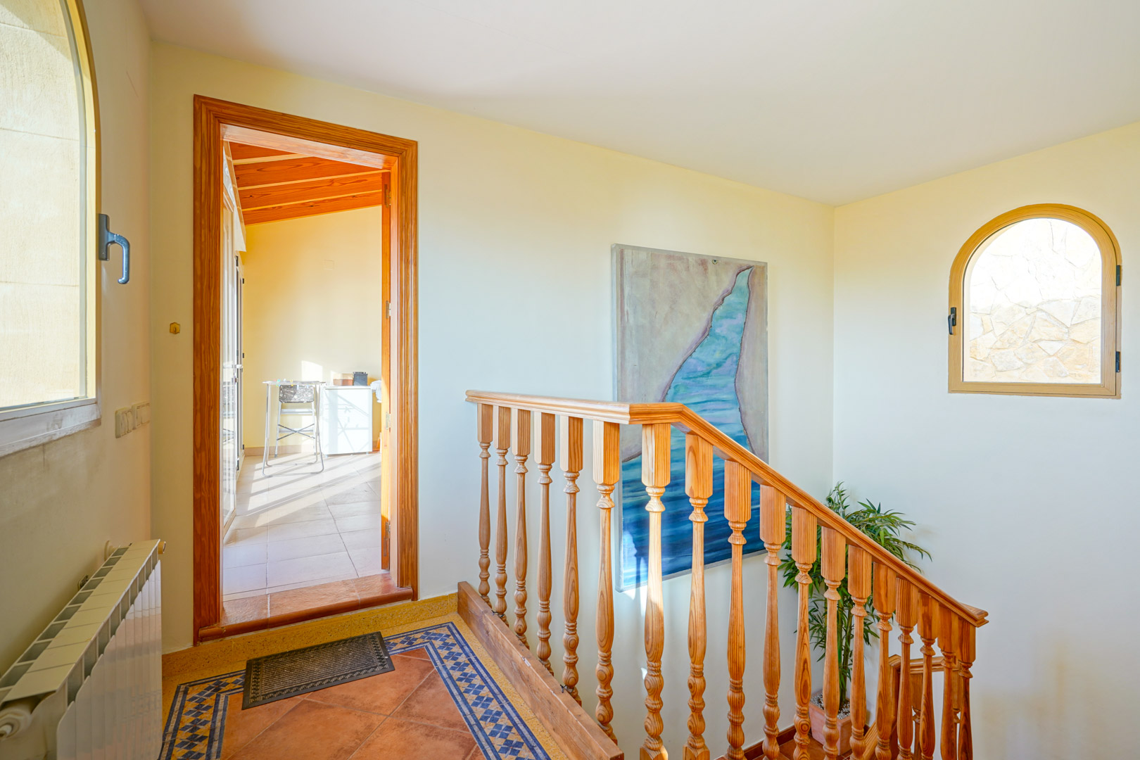Flur mit Holztreppe, Terrakottaboden mit dekorativen Bordüren und Rundbogenfenster. Ein Durchgang führt in einen hellen Raum mit sichtbaren Deckenbalken.