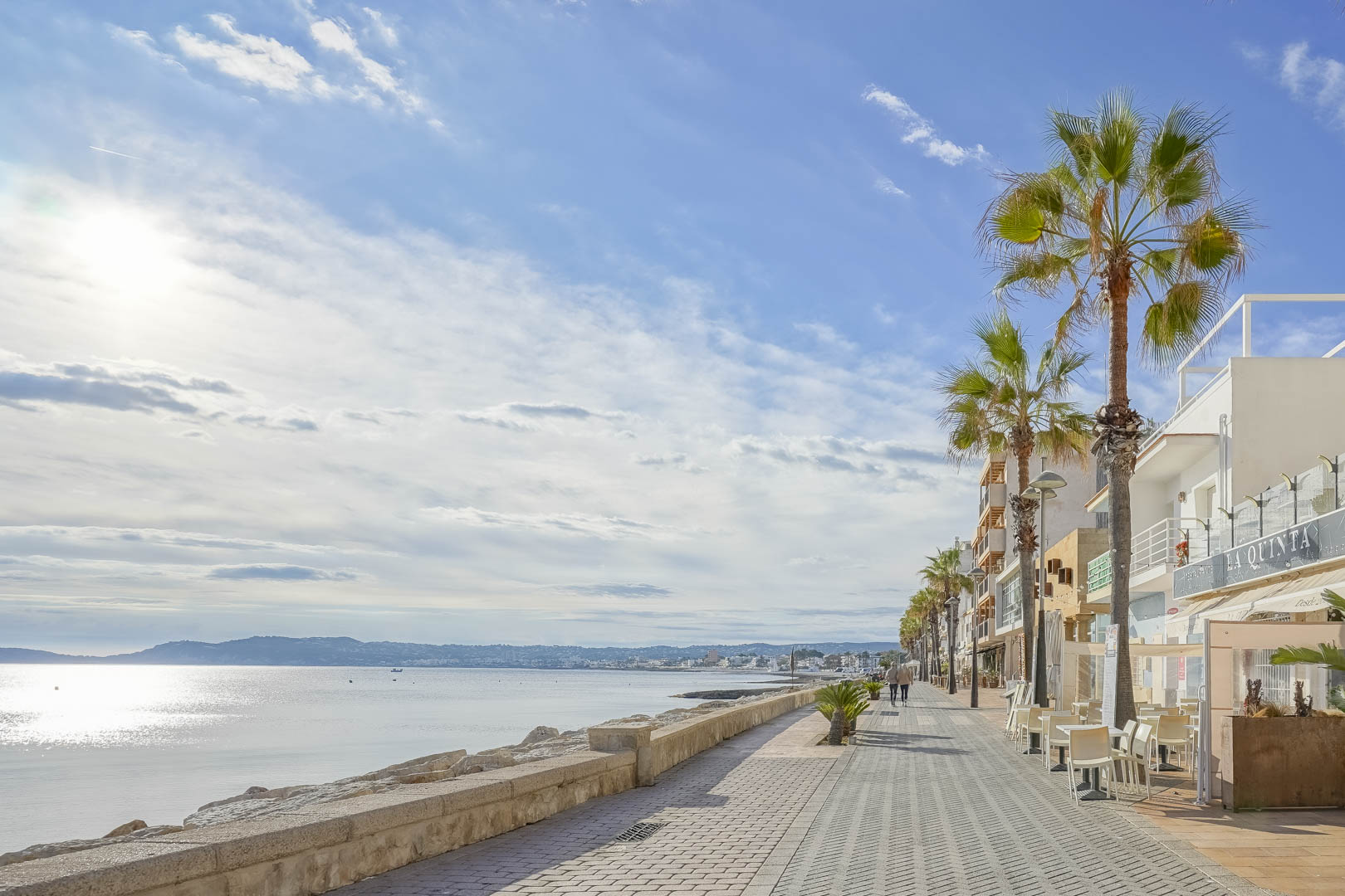 Küstenpromenade von Jávea mit Palmen, Straßencafés und Steinmauern, die einen weiten Blick über die Bucht und die Silhouette der Berge bietet.