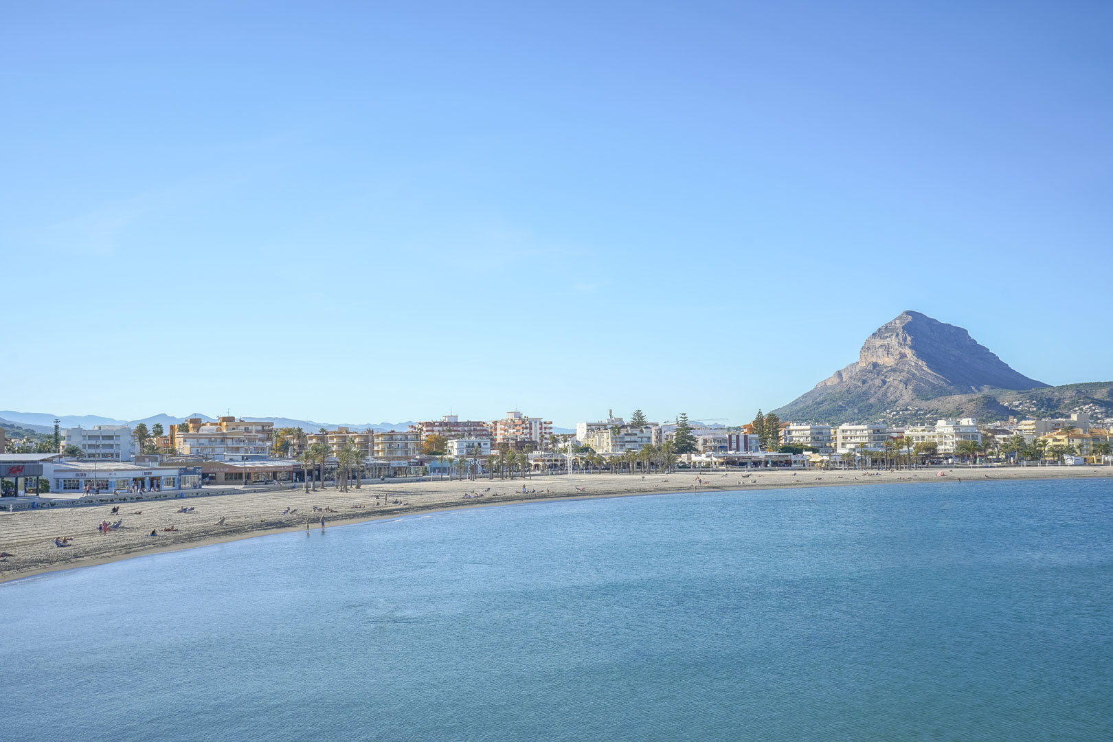Panoramablick auf die Bucht von Jávea mit dem weitläufigen Sandstrand, der Promenade und dem markanten Berg Montgó im Hintergrund.