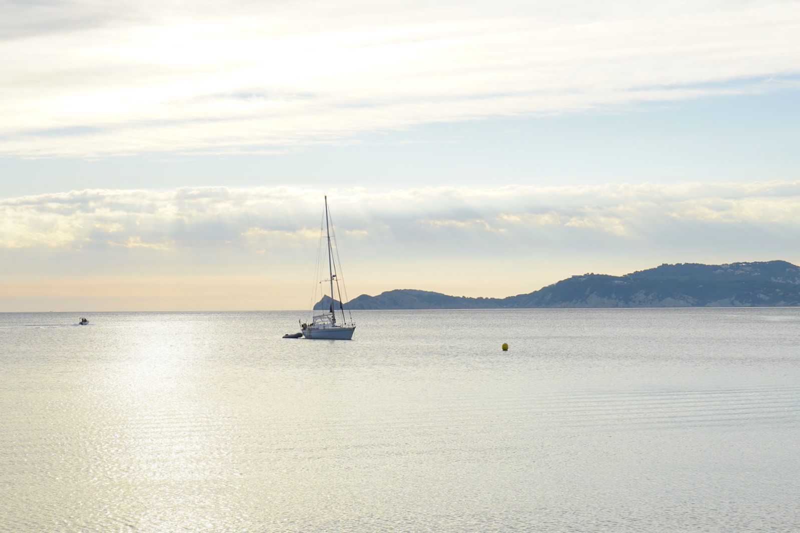 Panoramablick auf die Bucht von Jávea mit einem Segelboot auf ruhigem Wasser und der Silhouette des Cap Negre im sanften Morgenlicht.