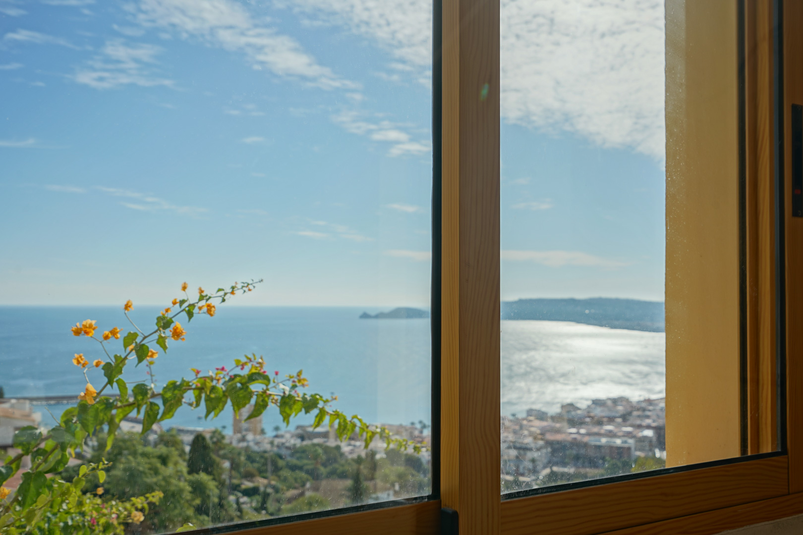 Panoramablick auf die Bucht von Jávea und das Cap Negre durch ein Holzfenster, akzentuiert durch blühende Bougainvillea.