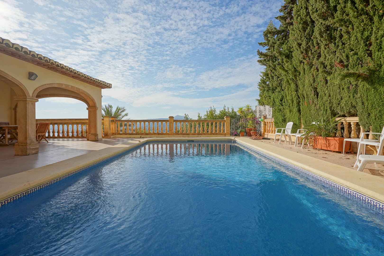 Rechteckiger Swimmingpool mit Steineinfassung, angrenzend an eine überdachte Terrasse mit Rundbögen und eine klassische Balustrade mit Meerblick.