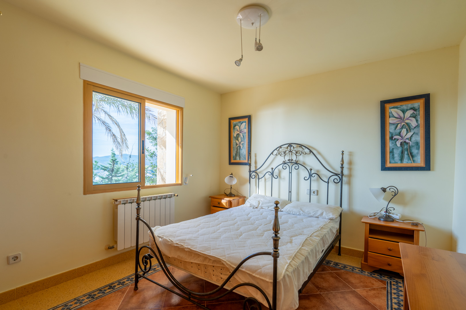 Schlafzimmer mit schmiedeeisernem Bettgestell, Terrakottaboden mit dekorativer Bordüre und Ausblick auf die mediterrane Landschaft.