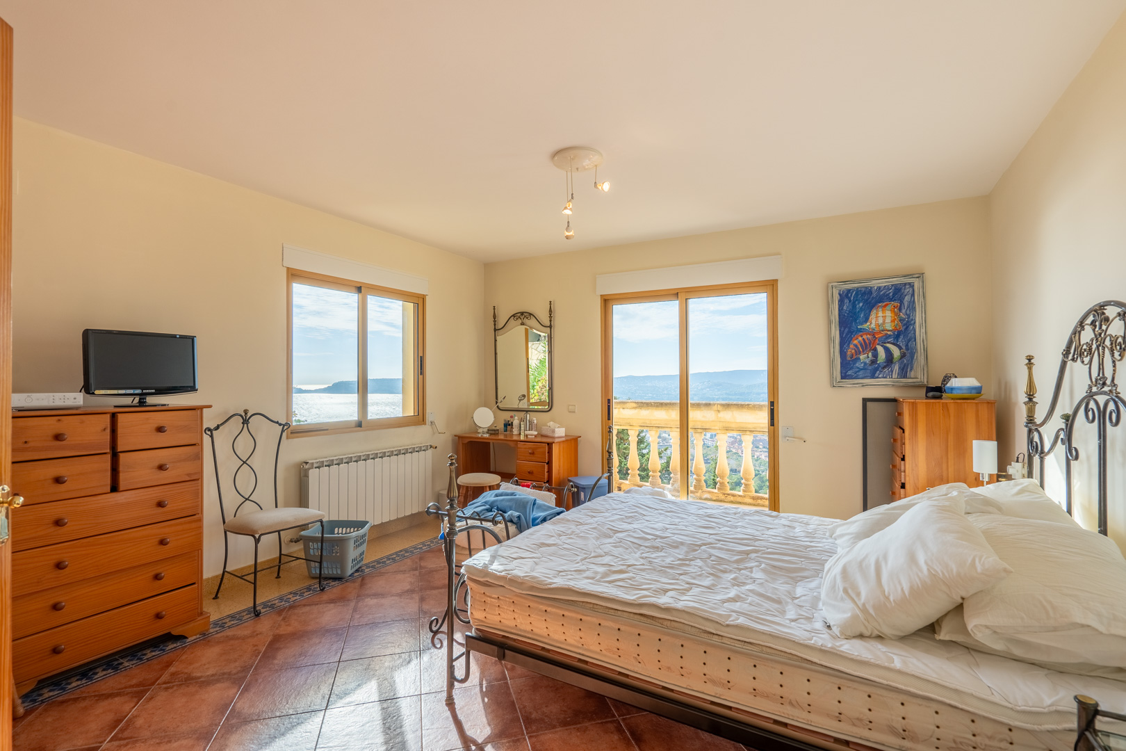 Schlafzimmer mit Terrakottaboden, Schmiedeeisenbett und Zugang zu einem Balkon mit Steinbalustrade, der einen weiten Blick auf das Meer und die Bucht bietet.