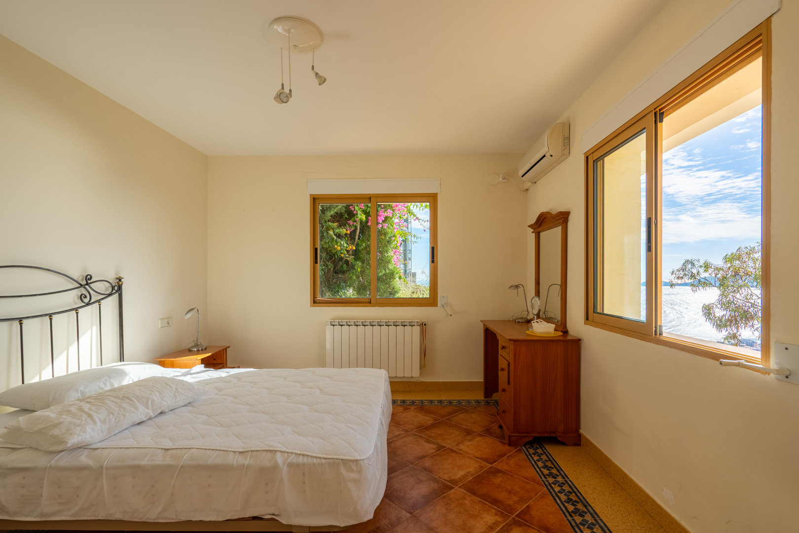 Schlafzimmer mit Terrakottafliesen und dekorativer Bordüre. Ausgestattet mit Holzfenstern mit Meerblick, einem schmiedeeisernen Kopfteil und Klimaanlage.