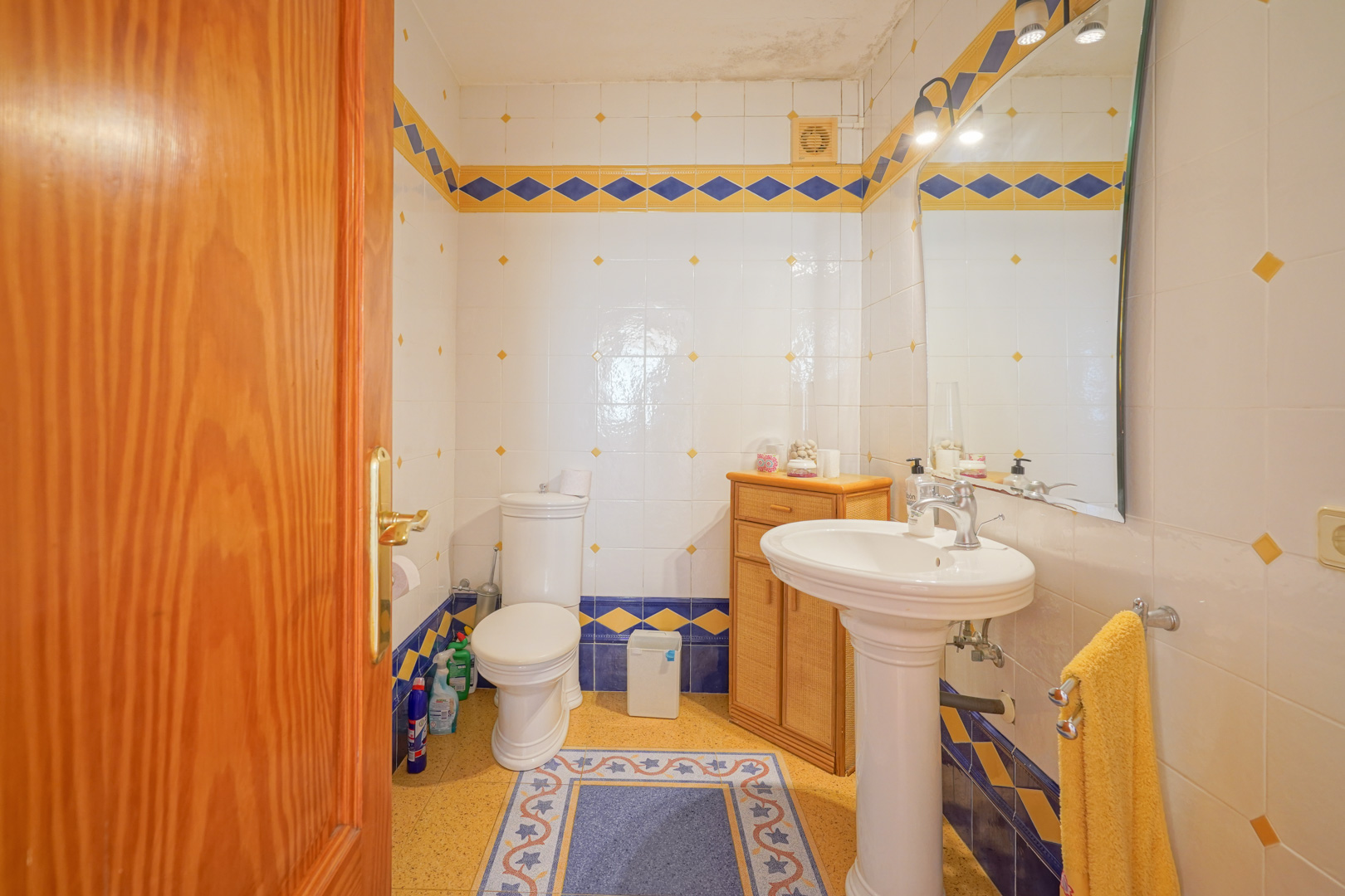 Traditionelles Badezimmer mit weißen Wandfliesen, blau-gelben Rautenmustern, einem Standwaschbecken und einem kleinen Holzschrank.