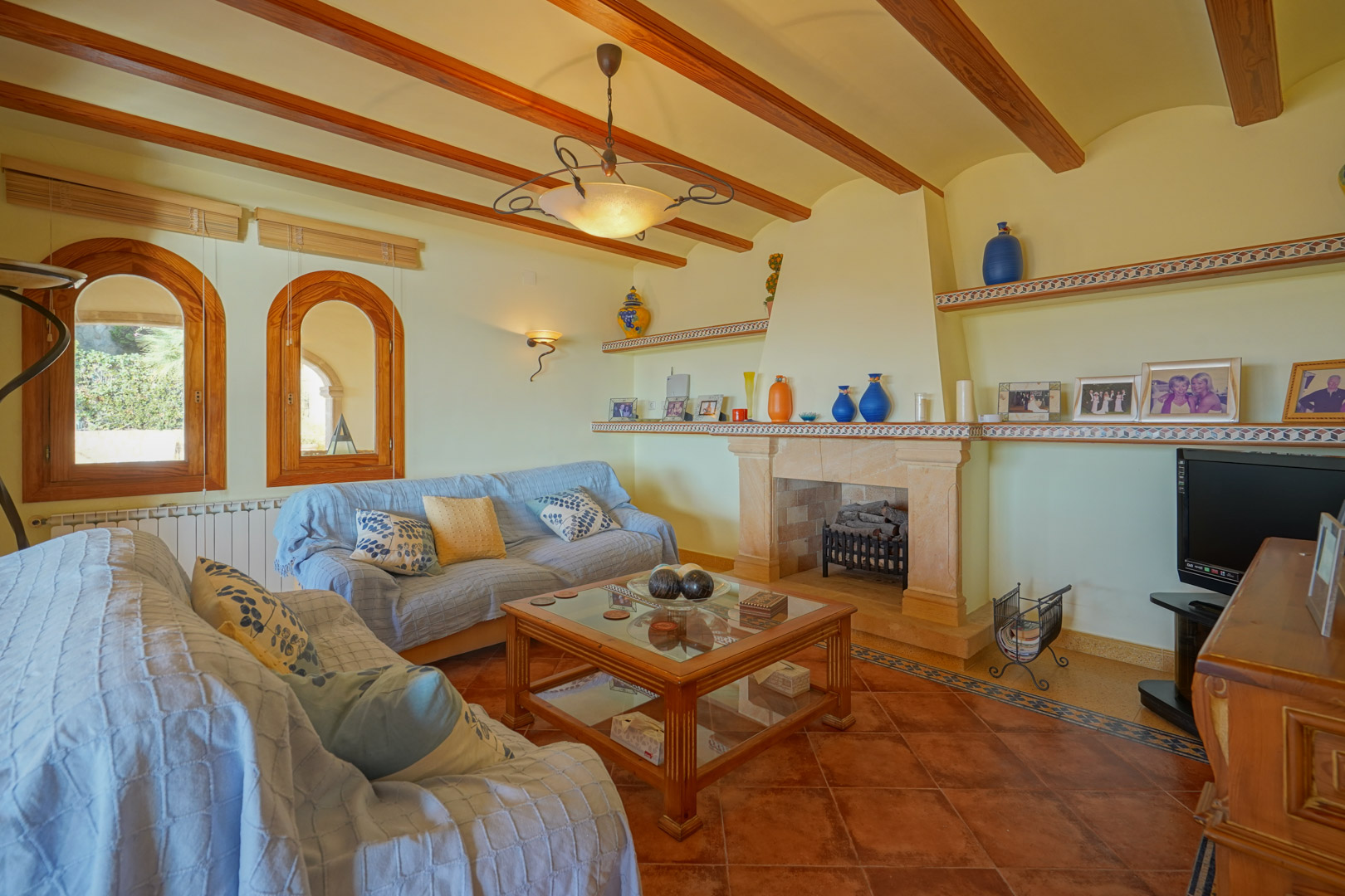 Traditionelles Wohnzimmer mit Steinkamin, sichtbaren Holzbalken und Terrakottaboden. Mit Rundbogenfenstern aus Holz und integrierten Regalen mit Keramikakzenten.