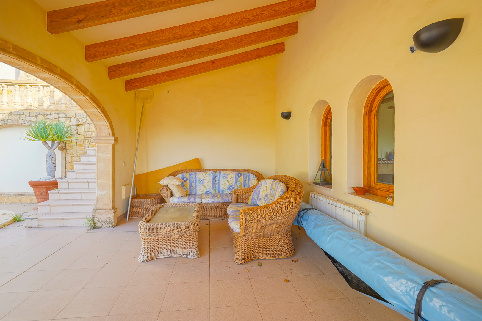Überdachte Terrasse mit sichtbaren Holzbalken, traditionellen Rundbogenfenstern und einer gemütlichen Rattan-Sitzgruppe im Außenbereich.