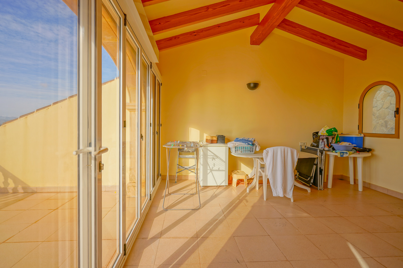 Verglaste Terrasse mit sichtbaren Deckenbalken aus Holz, Fliesenboden und großen Schiebefenstern für maximale Helligkeit.