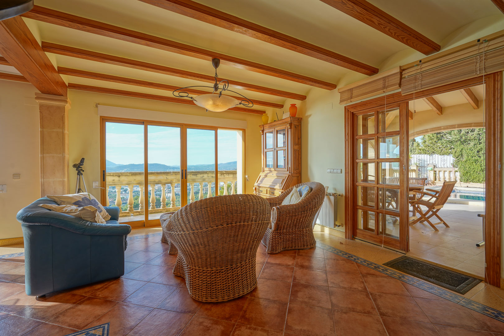Wohnzimmer mit Holzbalkendecke, Terrakottafliesen und Korbsesseln. Große Schiebetüren bieten einen weiten Blick auf das Tal und führen direkt zur angrenzenden Terrasse.